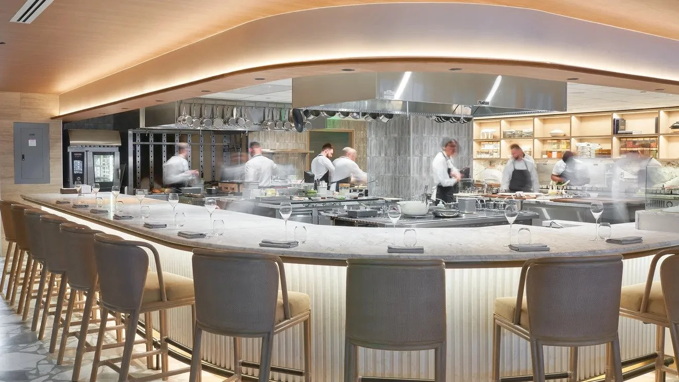 Modern restaurant kitchen with chefs preparing dishes, view from the bar seating area with empty glasses and napkins.