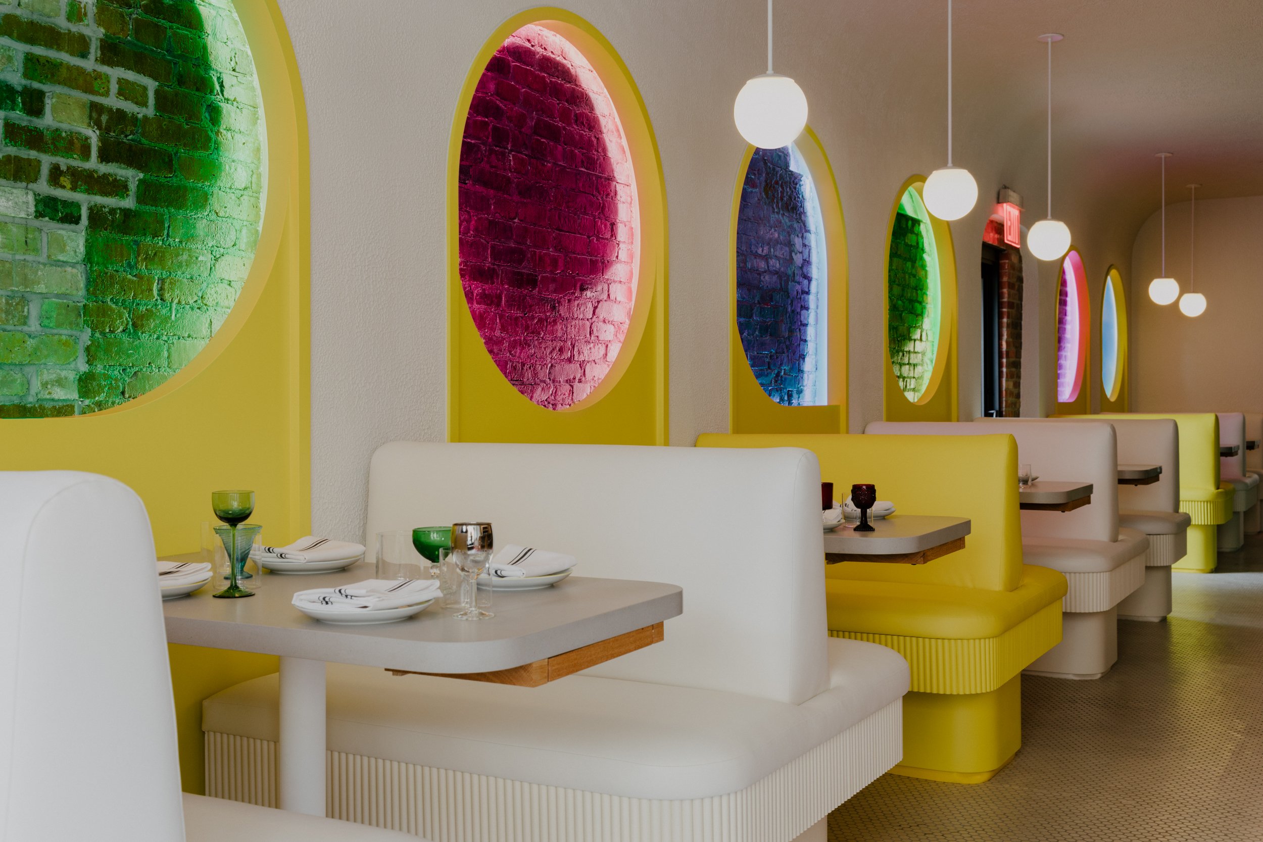 Empty restaurant booth seating with colorful, illuminated brick wall decor in green, pink, blue, and yellow. White and yellow upholstered booths, with set tables, in a modern dining interior.