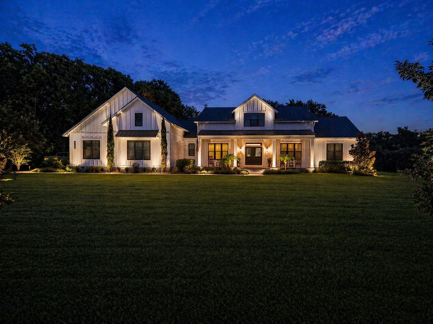 A large, modern house illuminated at night with exterior lights, surrounded by a well-maintained lawn and landscaped garden, under a dark blue sky.