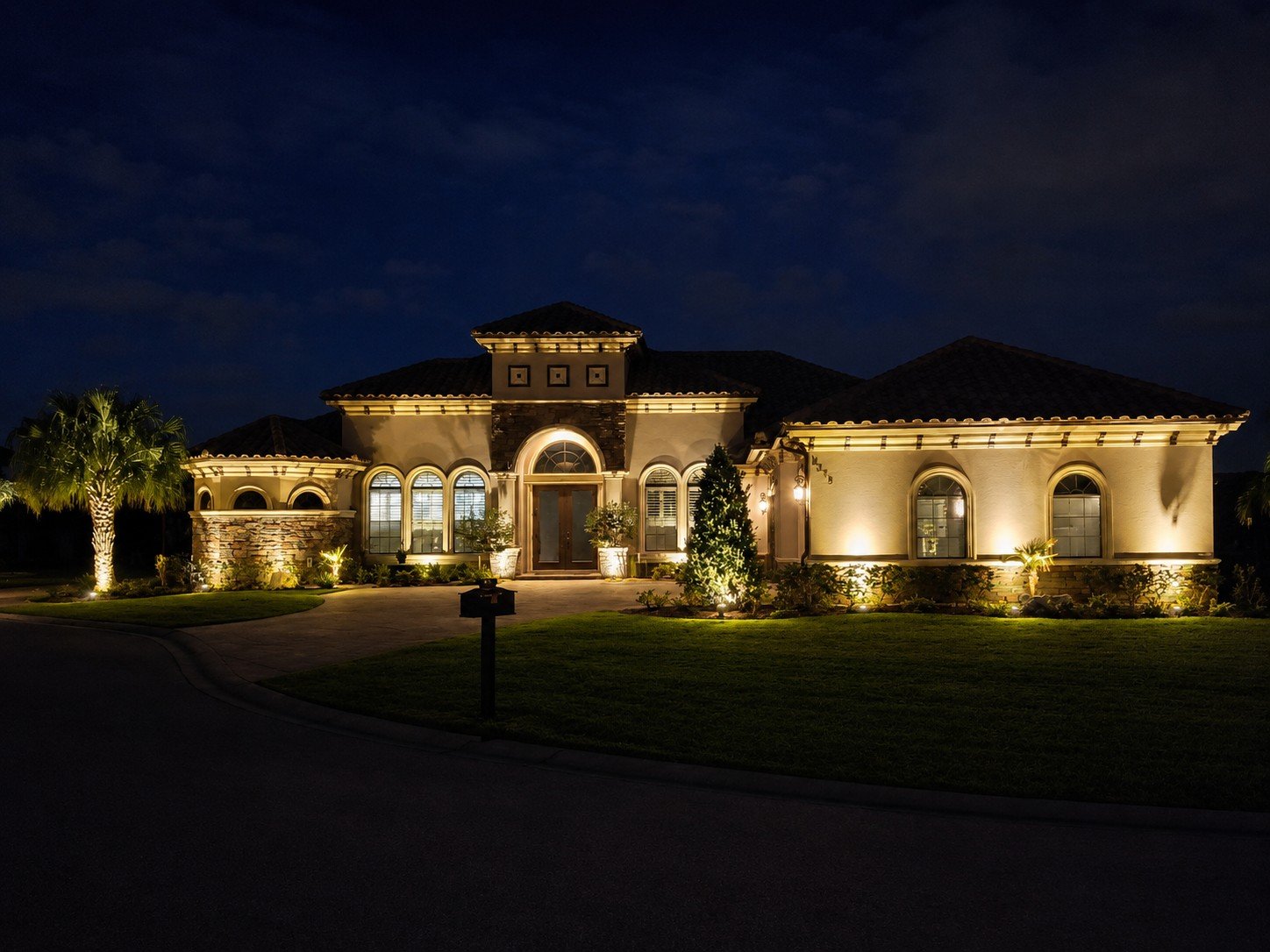 A large, elegant house at night with exterior lighting, palm trees, and landscaped front yard. Vibe Lighting Installation