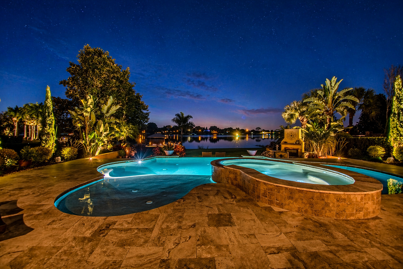 Night view of a backyard with a hot tub and pool, illuminated by outdoor lights, overlooking a lake with trees and houses in the distance, under a starry sky. Tampa Florida