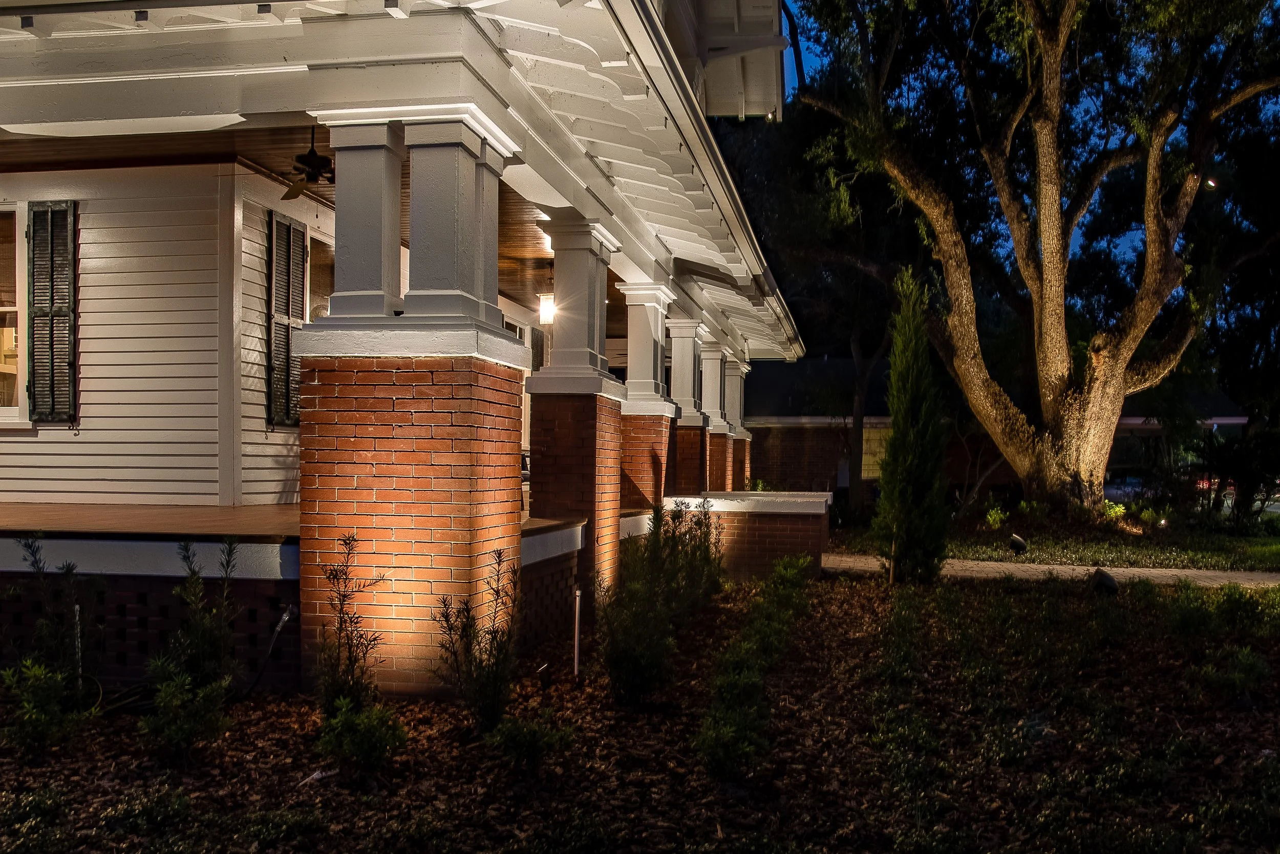 Nighttime view of a house with white siding, brick pillars, and a wrap-around porch by Vibe Lighting in Lakeland Florida. Landscape, outdoor, exterior lighting contractors.