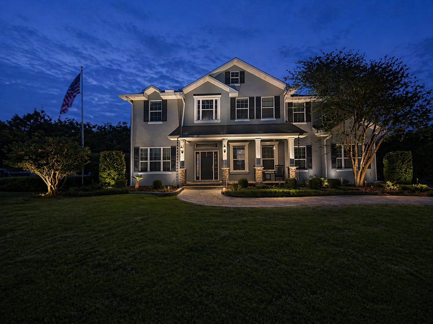A large, two-story modern house illuminated at night with outdoor lighting, surrounded by well-kept lawn and bushes, with an American flag on a flagpole to the left. Clearwater FL