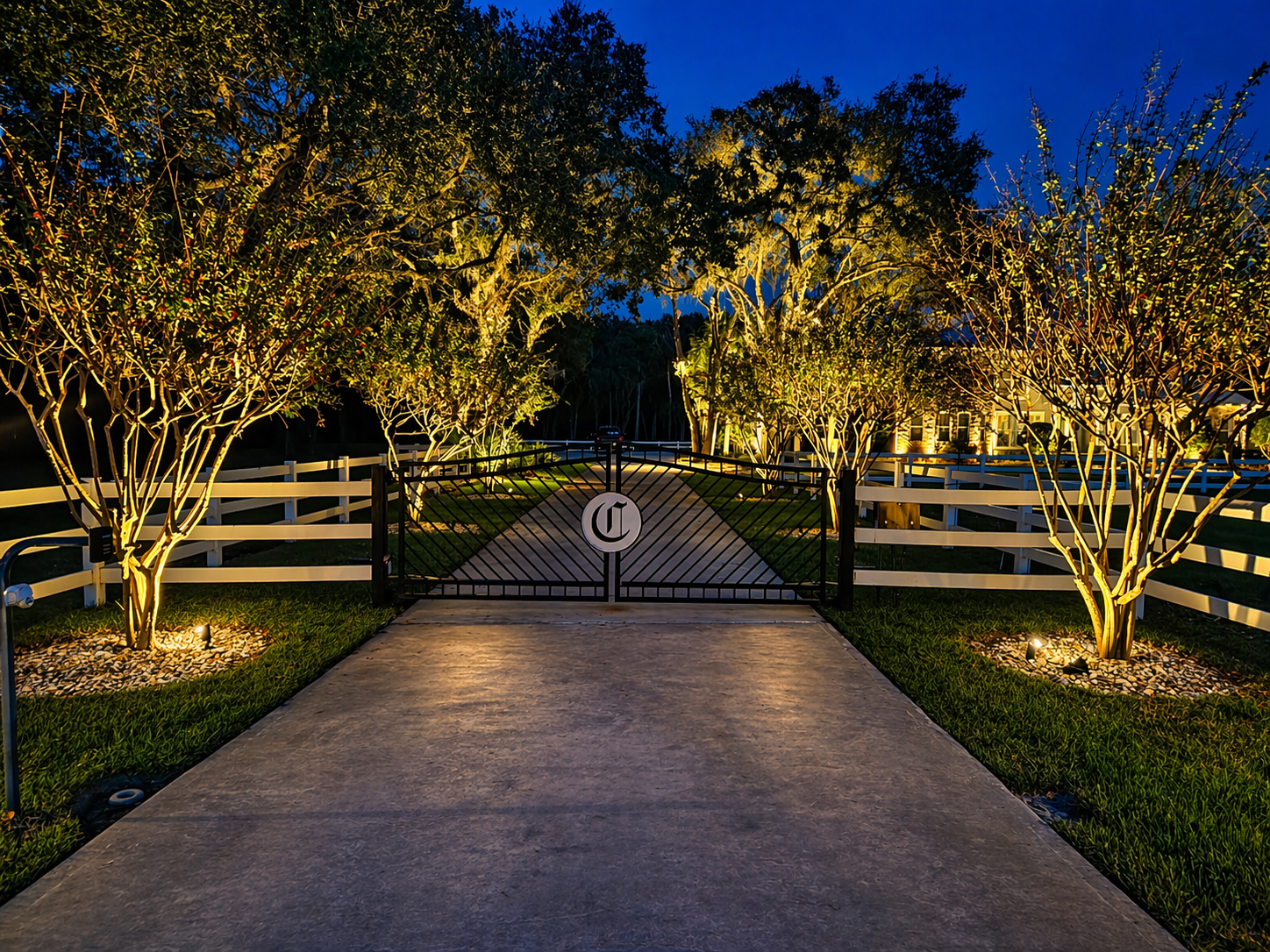 A gated entrance with a logo, surrounded by illuminated trees and a white fence, at dusk. Polk County Florida