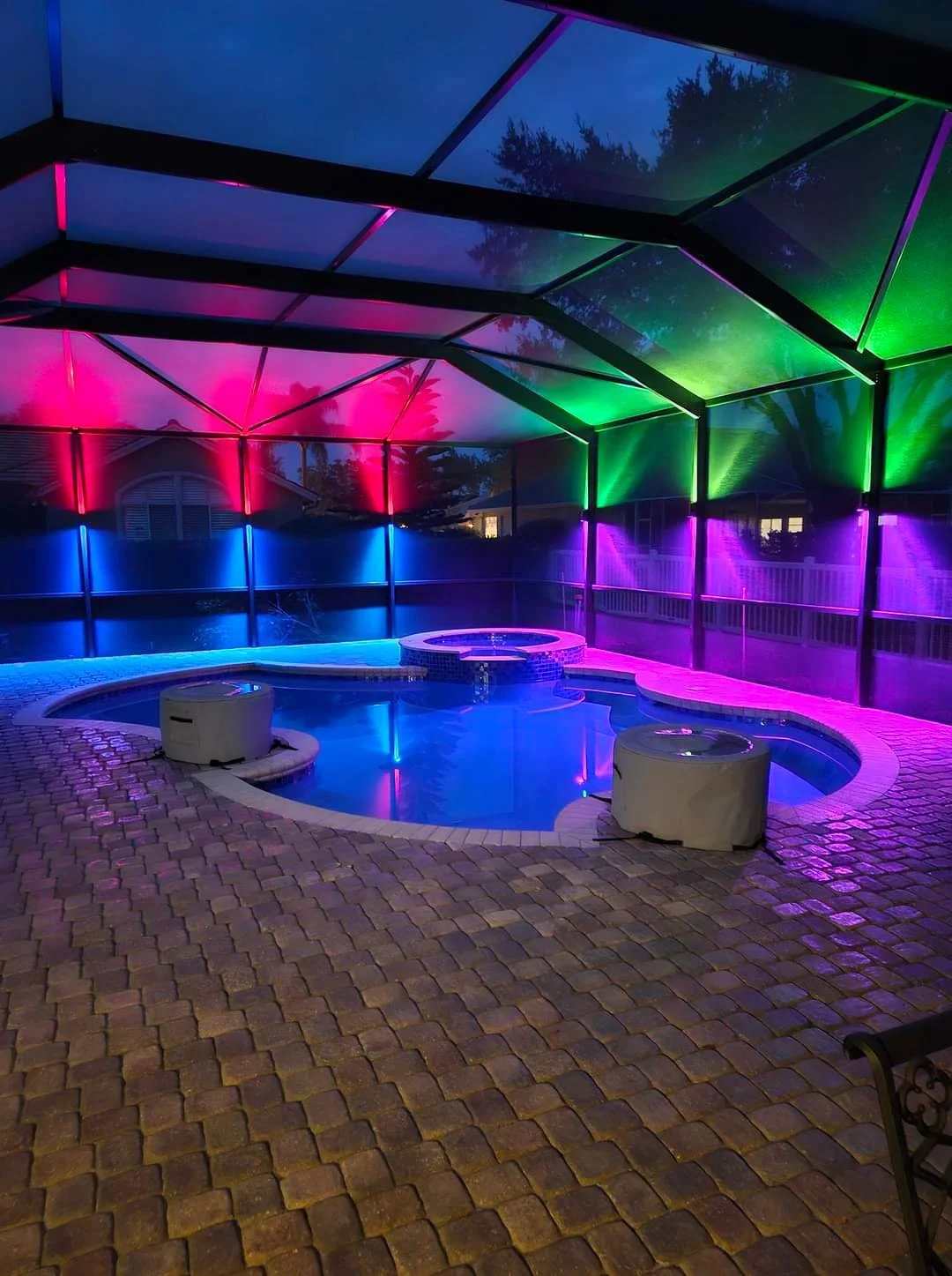 Nighttime view of a swimming pool enclosed by a screen with colorful LED lights illuminating the enclosure by Vibe Lighting in Lakeland Florida. Landscape, outdoor lanai lighting.
