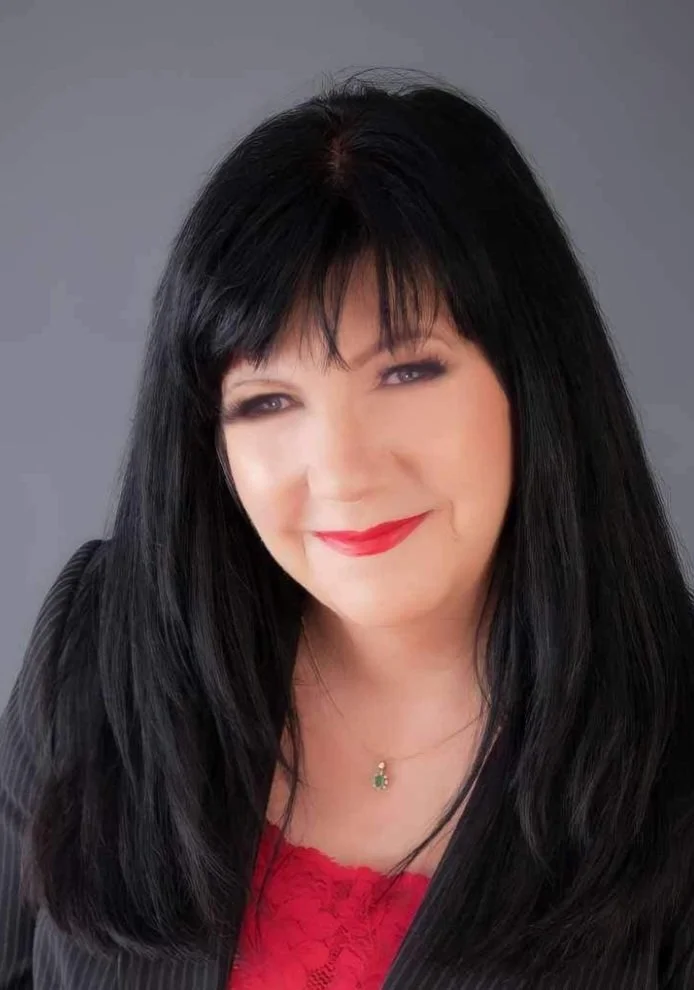 A woman with long black hair, wearing a black blazer over a red lace top, smiling with red lipstick, and a small necklace with a green pendant, against a grey background.