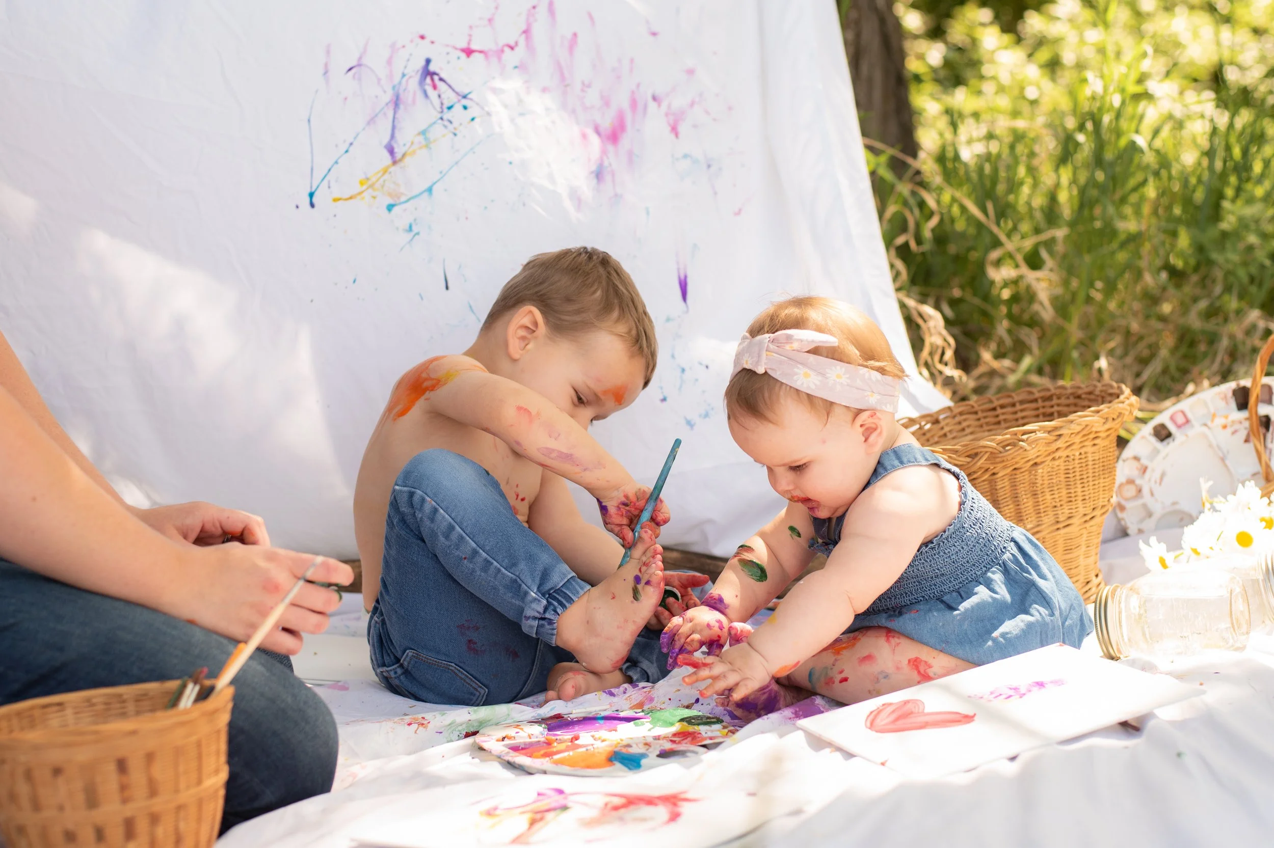 Two young children, a boy and a girl, are playing with finger paints outdoors on a sheet of paper. The children are covered in colorful paint and seem to be enjoying creative art activities.