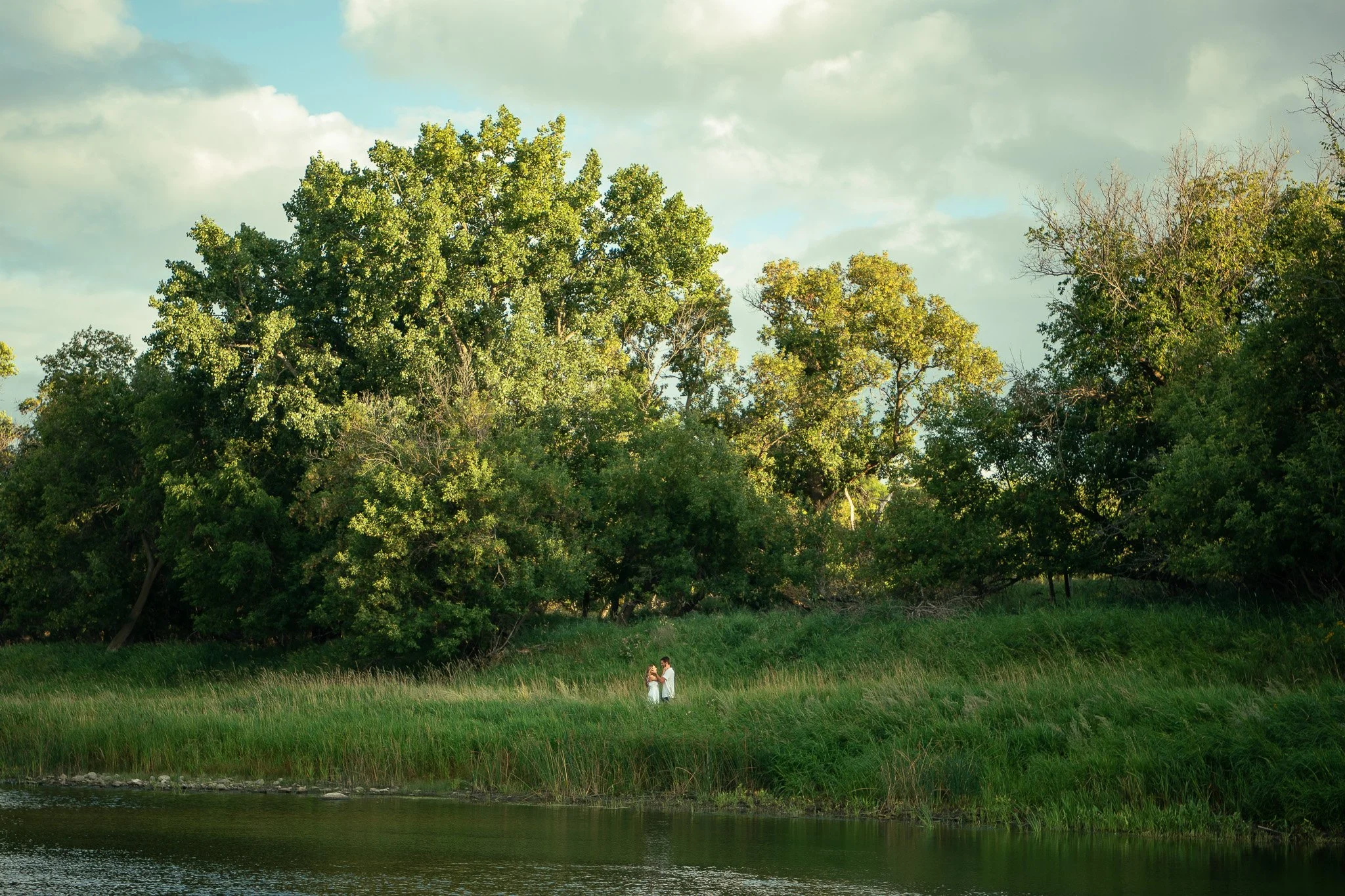A couple standing on grassy riverbank, surrounded by trees with green foliage, under a partly cloudy sky.