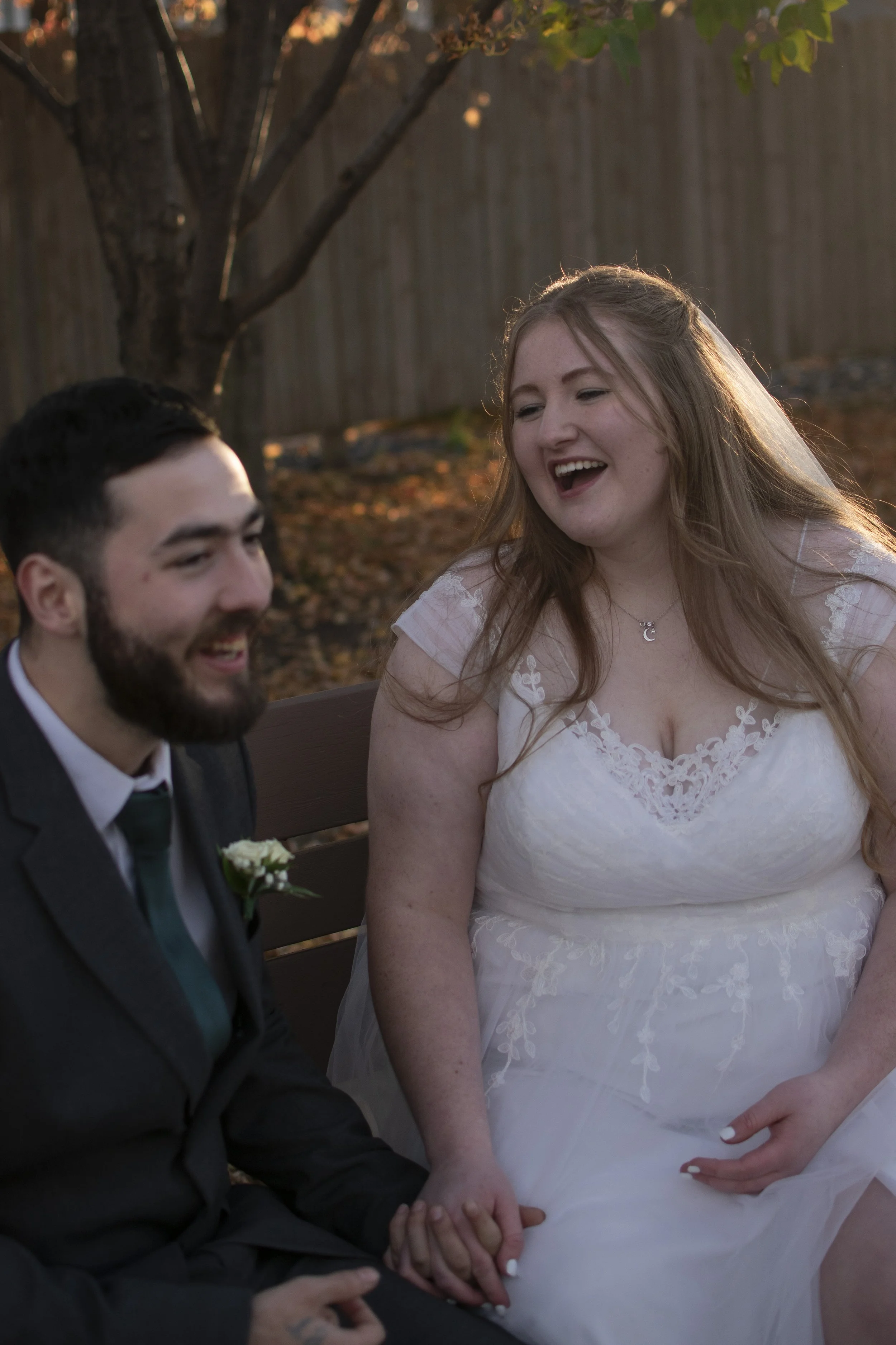 A couple, a man with a beard and a woman with long hair in a wedding dress, holding hands and sitting on a bench outdoors during sunset.