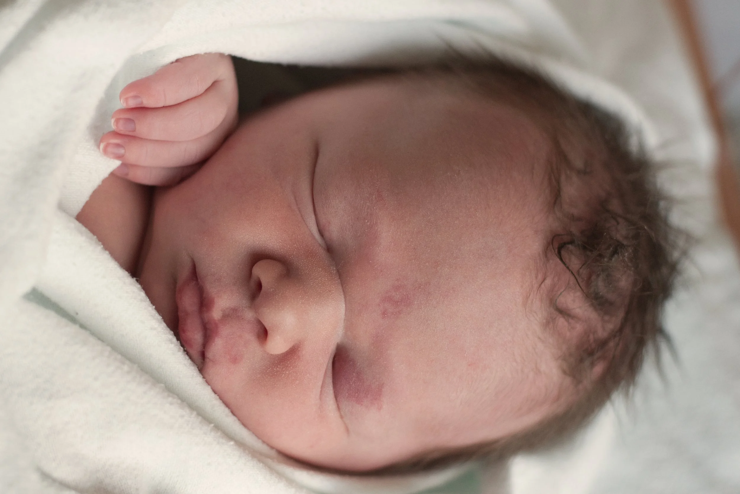Close-up of a sleeping newborn baby with hand resting near face, wrapped in a white blanket.