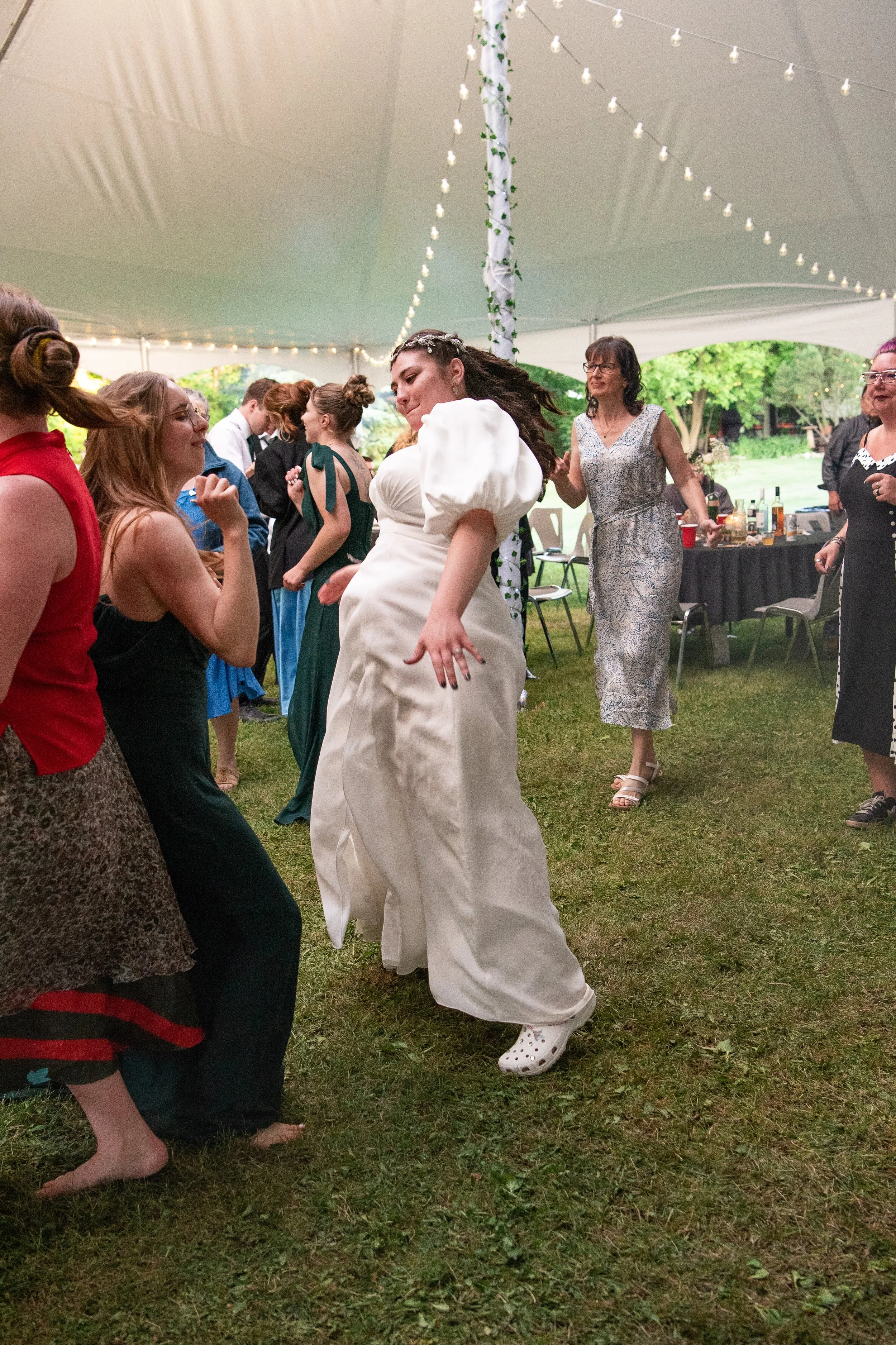 People dancing at a celebration under a tent decorated with string lights, with grass on the ground and a table with drinks in the background.