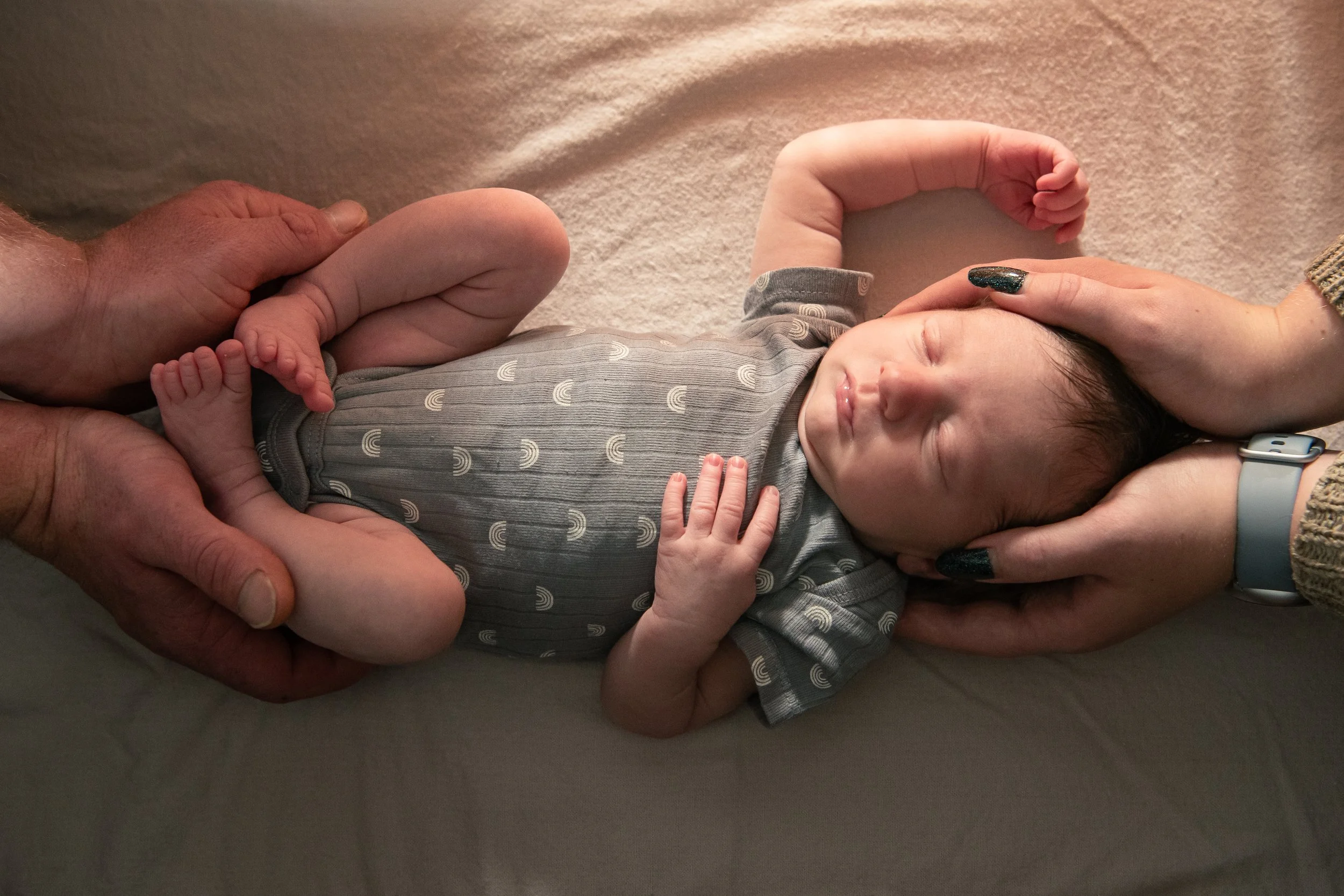 A newborn baby lying on a soft surface, being gently held by two adults. The baby is peacefully sleeping, wearing a gray onesie with a rainbow pattern. One adult is cradling the baby's head and the other is supporting the baby's legs.