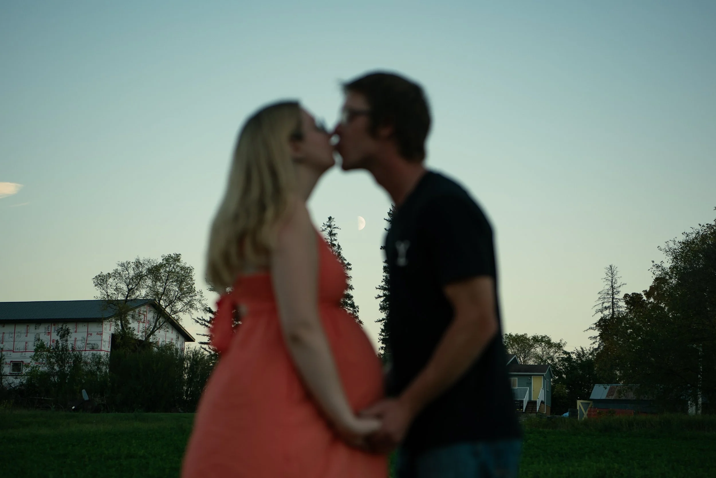 A blurry outdoor scene of a man and woman kissing, holding hands, with a house, trees, and a visible moon in the sky.