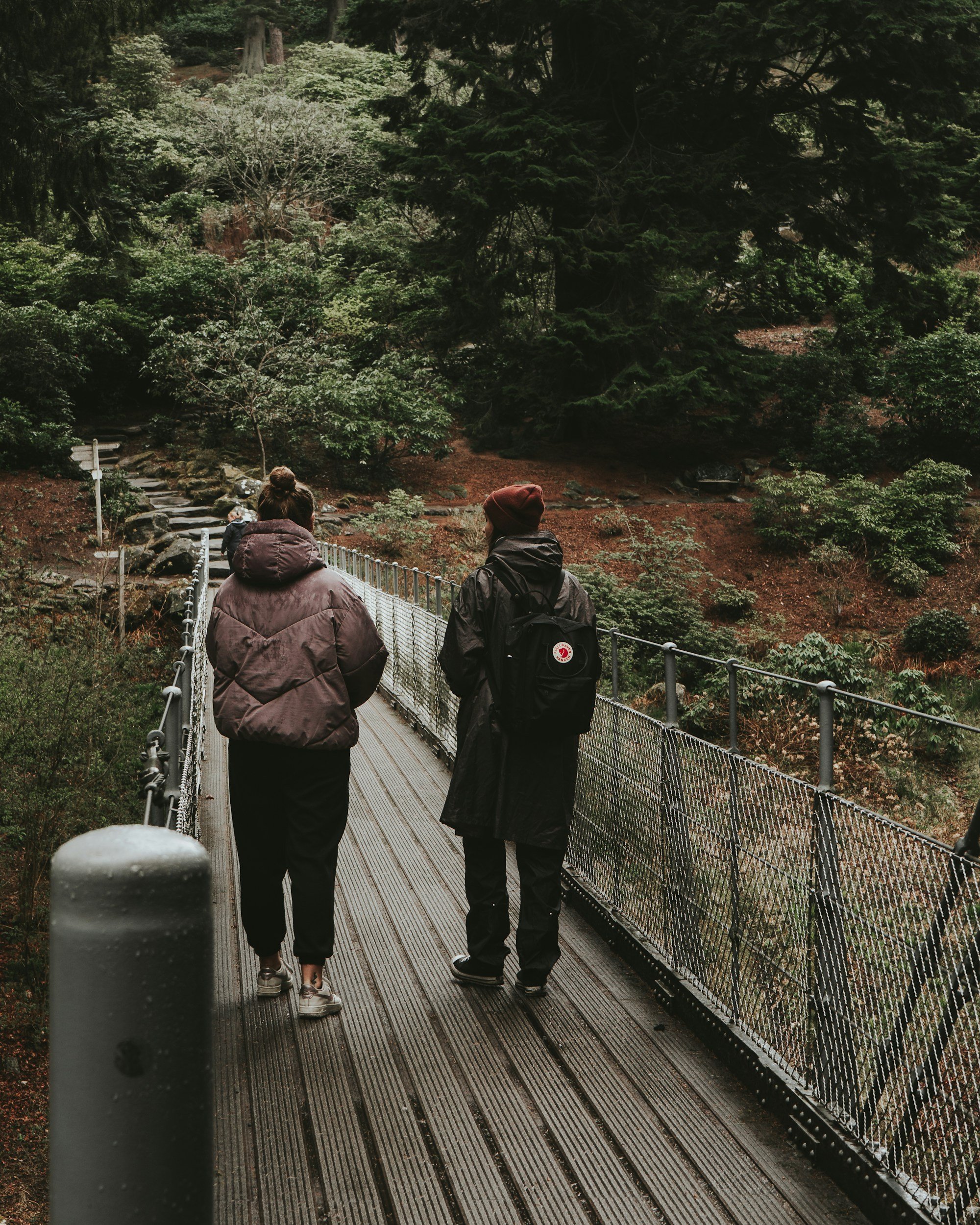 Two women walking on a narrow suspension bridge surrounded by lush green trees and foliage in a forest setting.