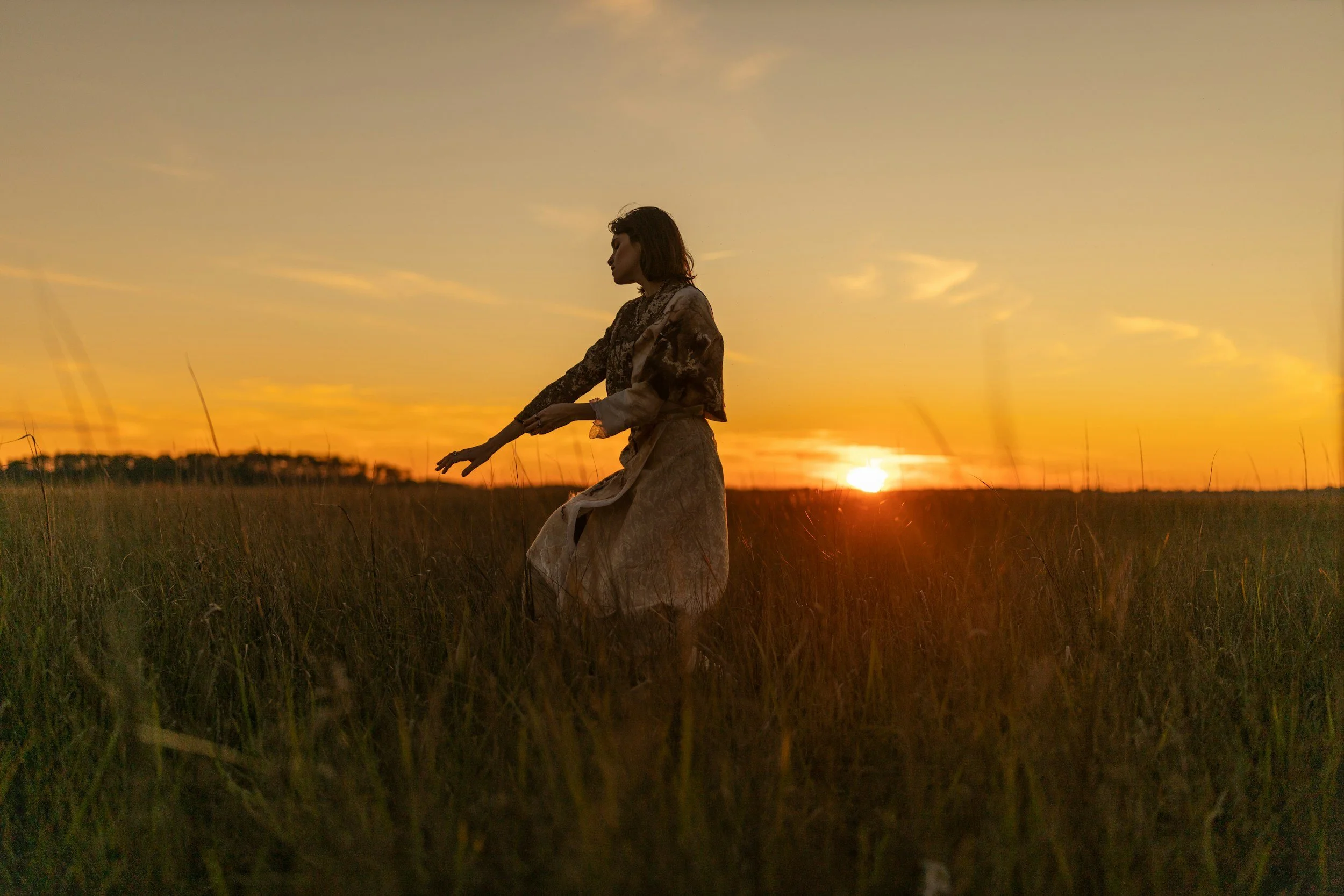 A woman standing in a field at sunset with her arms outstretched and a silhouette against the golden sky.