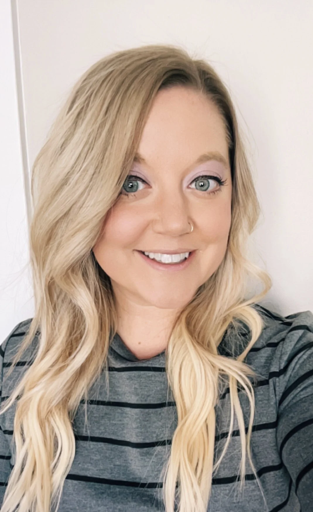 A smiling woman with long, wavy blonde hair, blue eyes, light makeup, wearing a gray and black striped shirt, standing against a white background.