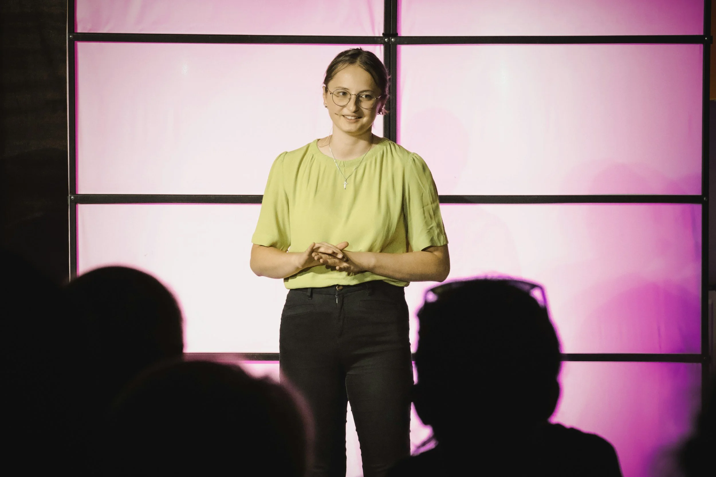 A woman with glasses, wearing a light green blouse and black pants, speaking on stage with a pink and white background behind her, and an audience silhouette in the foreground.