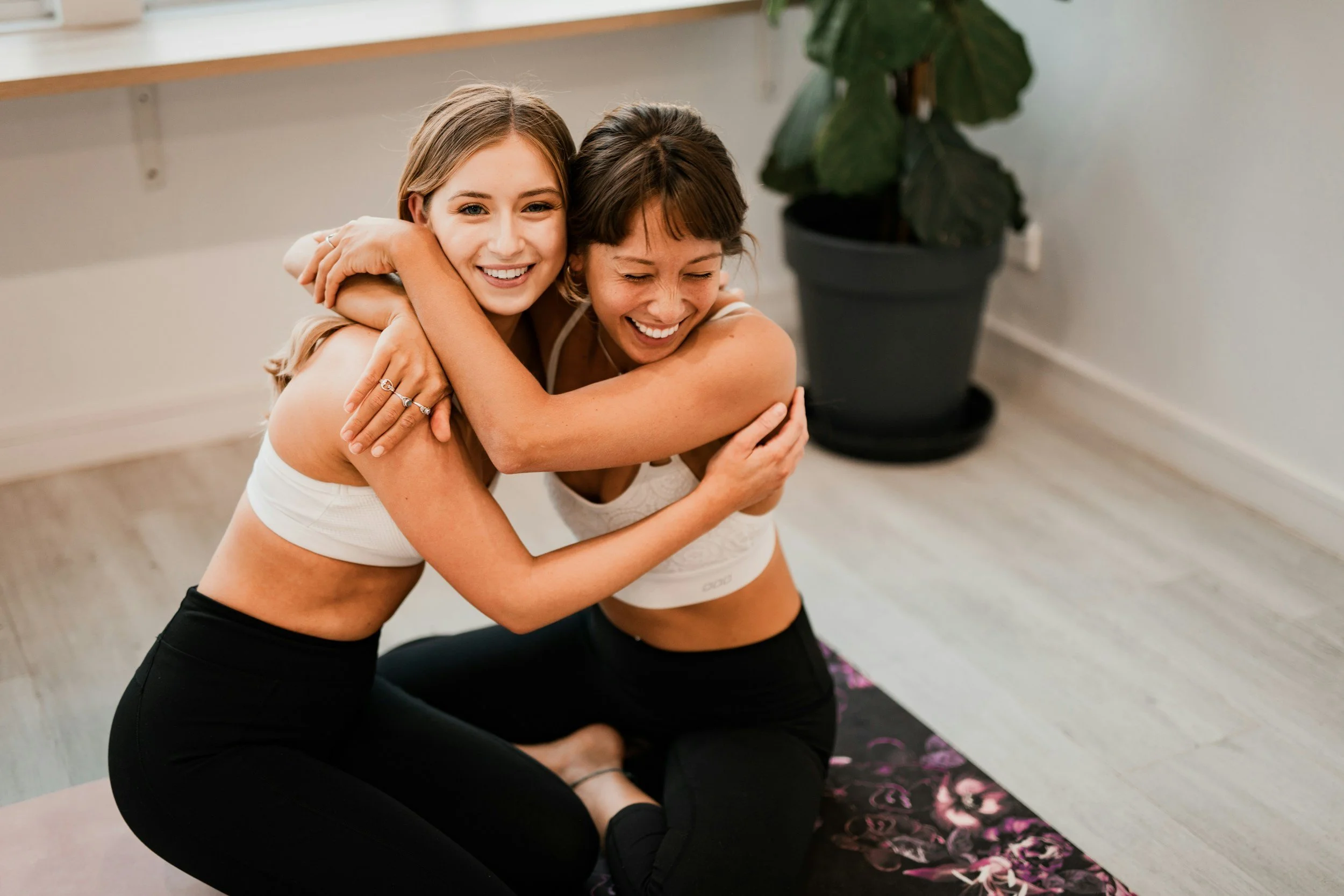 Two women practicing partner yoga or stretching together indoors, hugging each other around the shoulders with their heads close together and smiling.