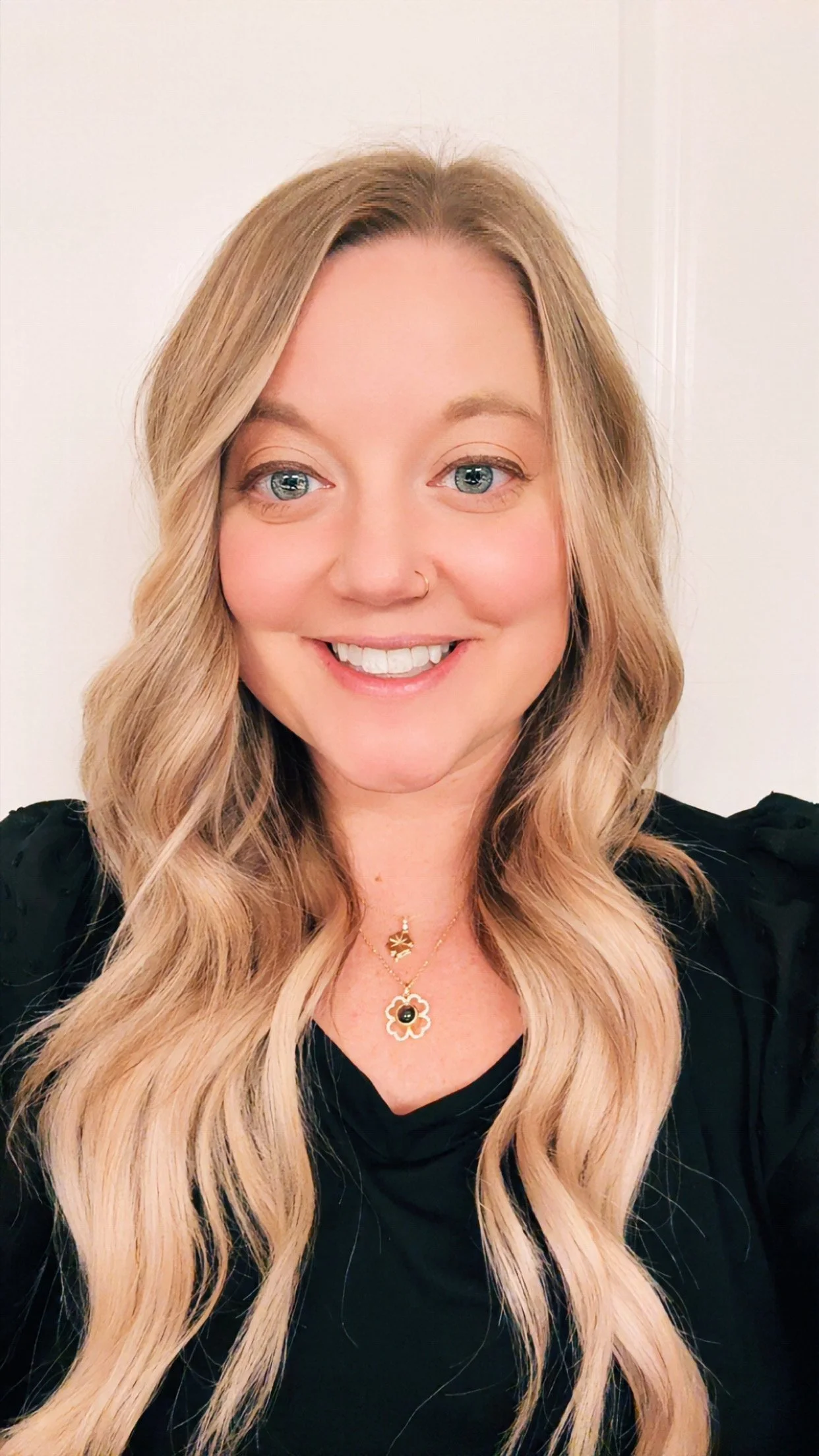 Close-up of a woman with long, wavy blonde hair, blue eyes, light makeup, a nose piercing, smiling, wearing a striped shirt.