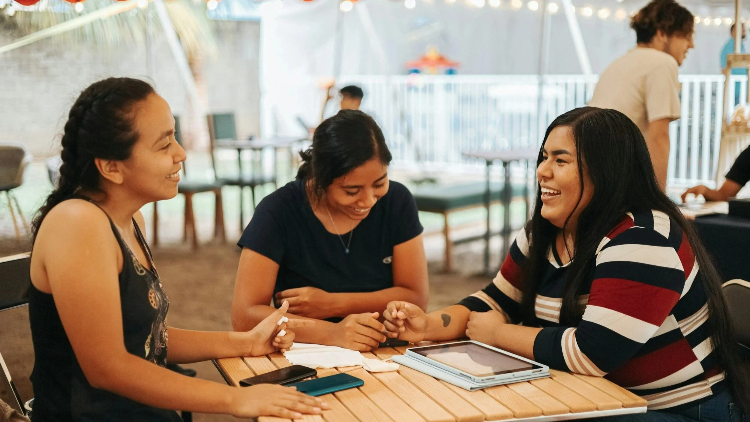Three women laughing and holding hands at a table in a casual indoor setting, with a fourth person in the background.