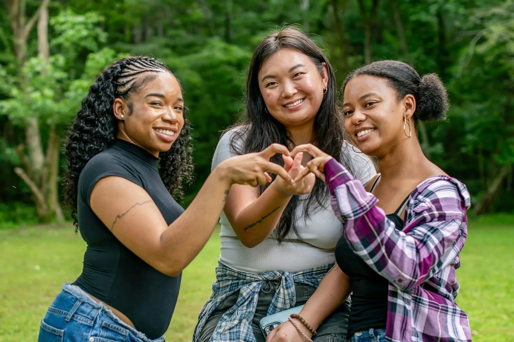 Three women standing outdoors in a wooded area making a heart shape with their hands, smiling.