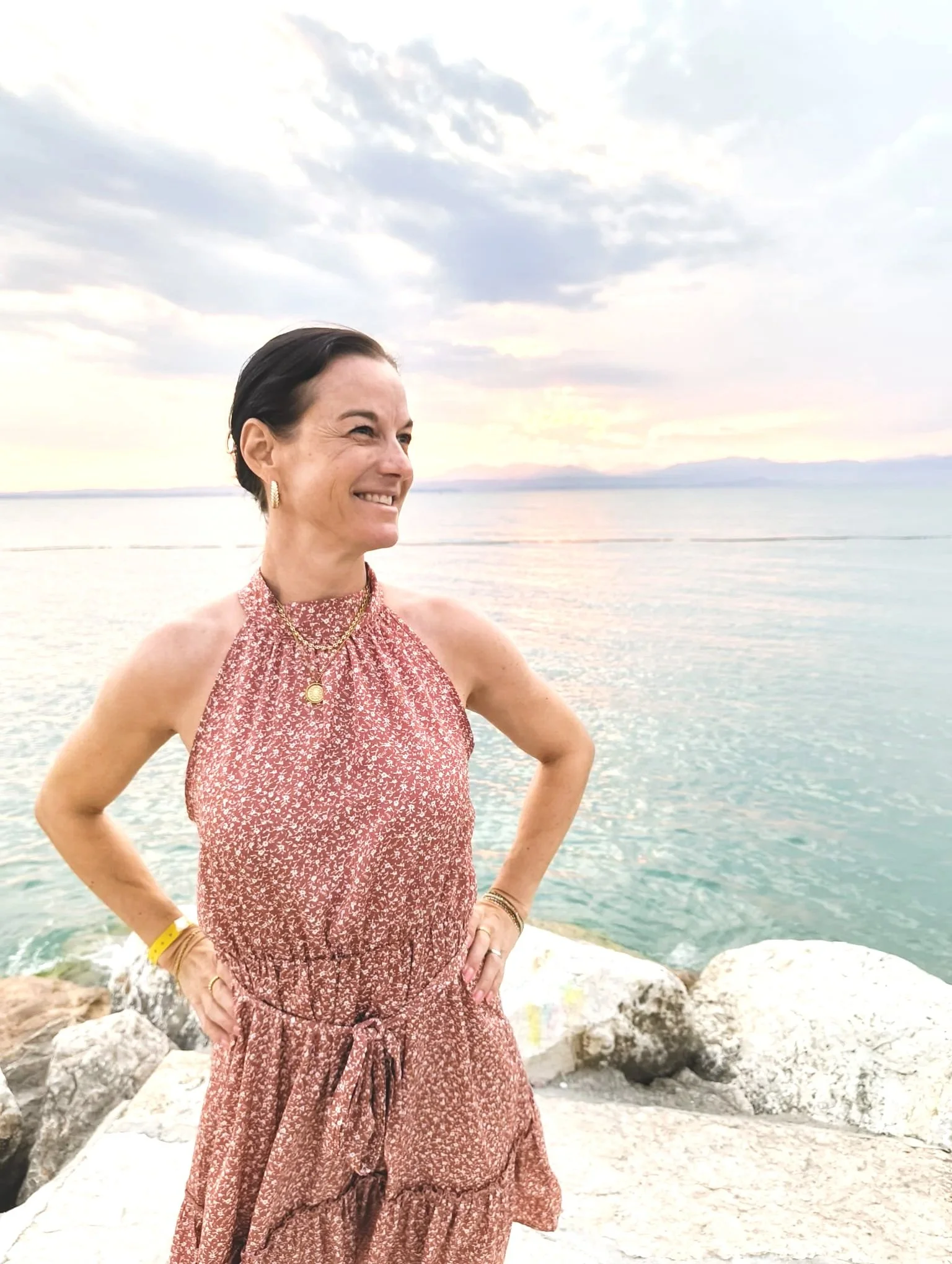 Eine lachende Frau in einem roten Blumenmuster-Kleid steht am Strand mit Steinen, das Meer und ein Himmel mit Wolken im Hintergrund, es ist Sonnenuntergang.