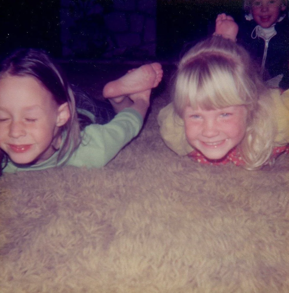 Two children lying face down on a carpeted floor, with one child's arm resting on the other, smiling and holding hands, in a cozy indoor setting.