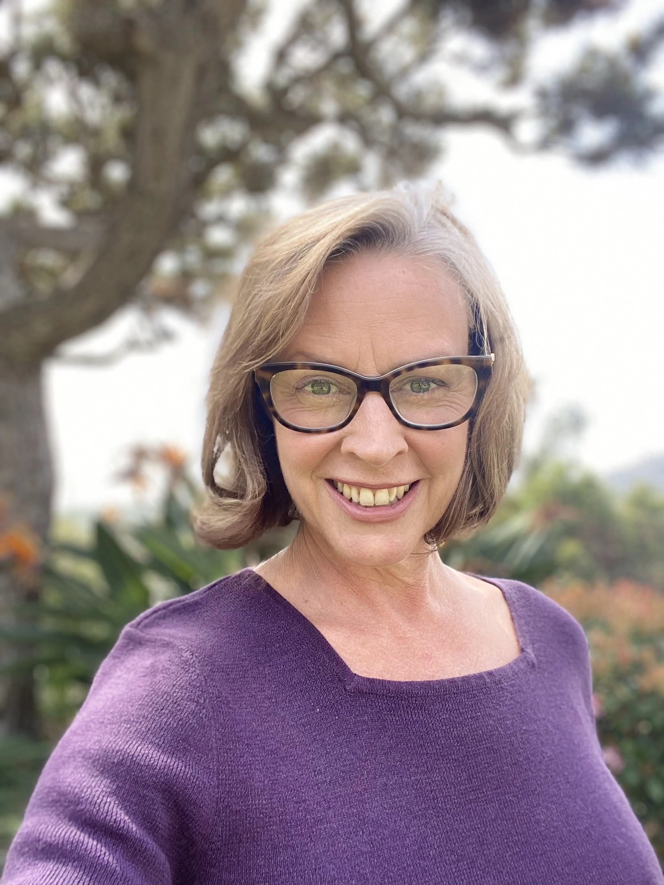 A smiling woman with glasses standing outdoors, wearing a purple top, with a tree and greenery in the background.