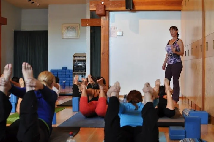 A group of adults participating in a yoga class indoors, lying on mats with their legs raised and hands supporting their feet, while an instructor stands nearby.