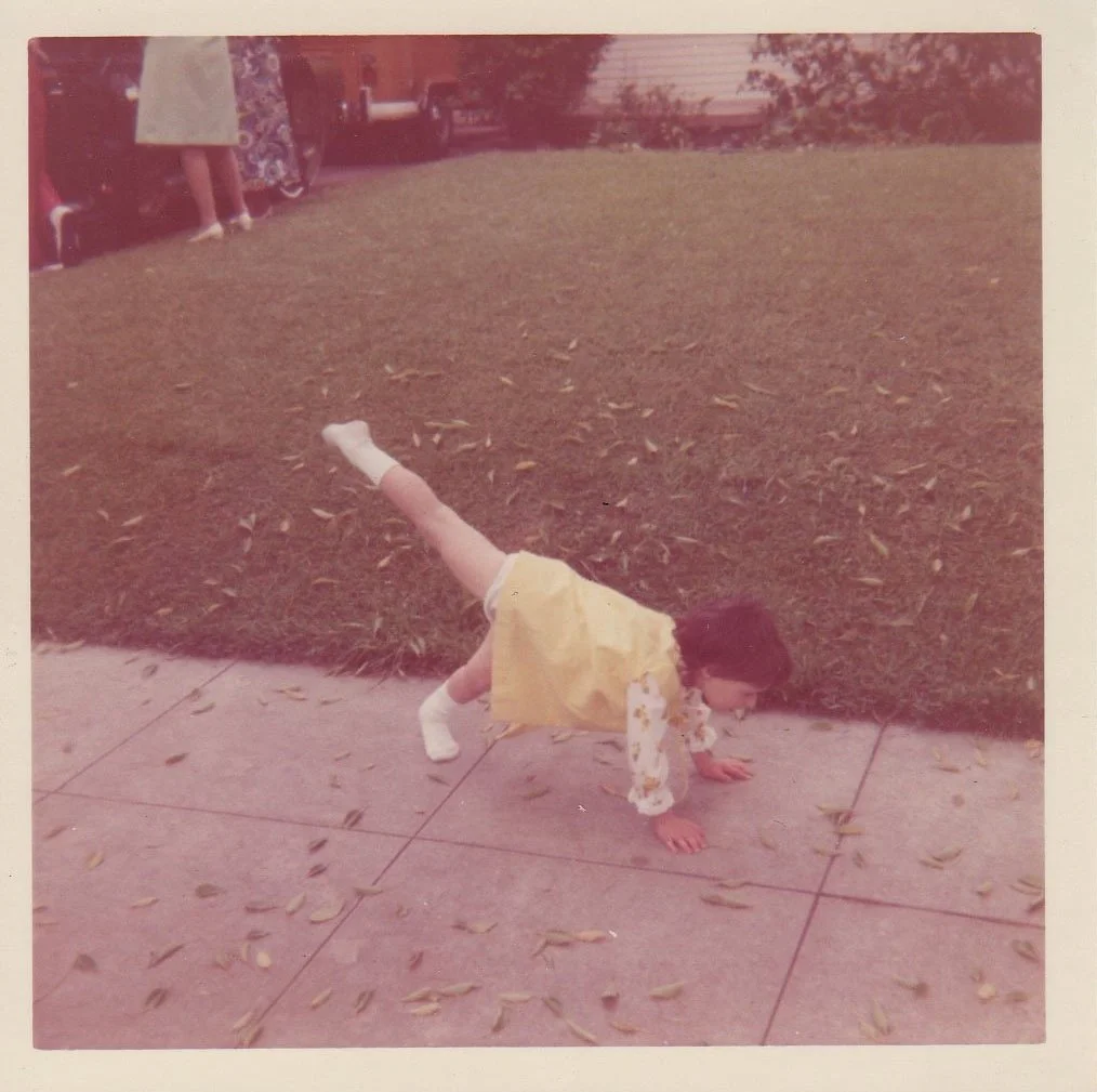 A young girl performing a handstand on a sidewalk with fallen leaves, wearing a yellow skirt, floral long-sleeve shirt, and white socks, with people and trees in the background.