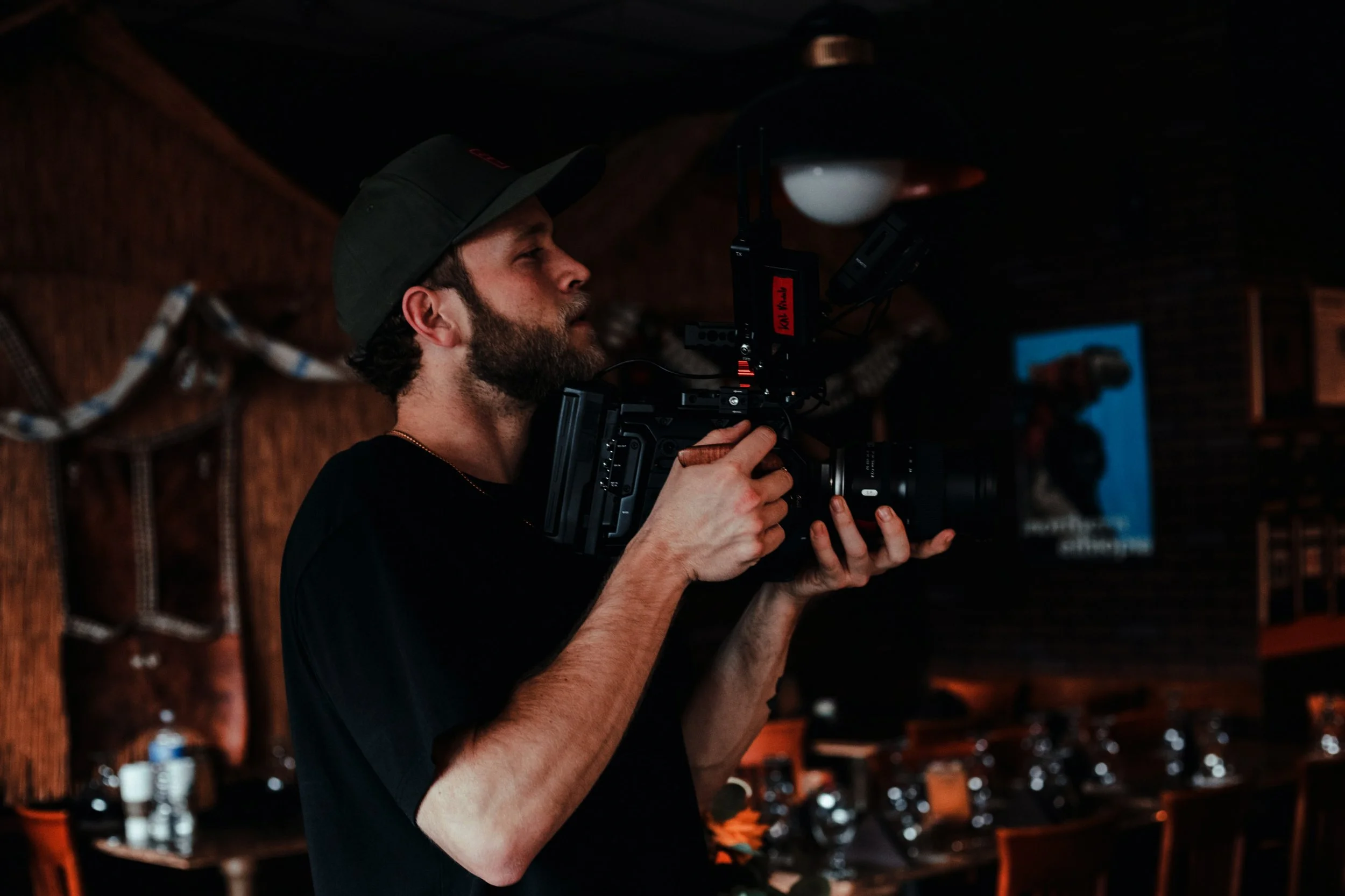 A man with a beard and dark cap operating a professional video camera indoors at a dimly lit venue.