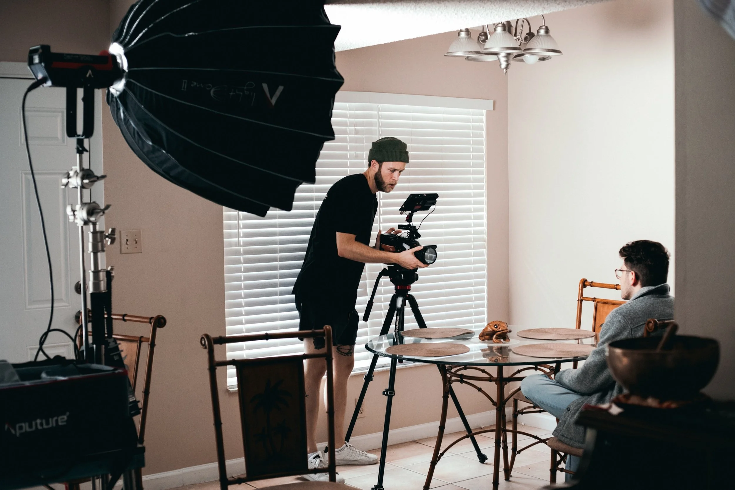 A man is filming a person sitting at a dining table with a professional video camera and lighting equipment inside a house.