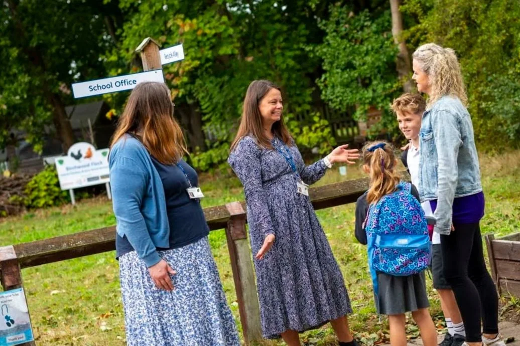 A group of five people, including children and adults, standing outdoors near a wooden fence, engaged in conversation. There are signs in the background indicating a school and a nature area, with trees and greenery around.