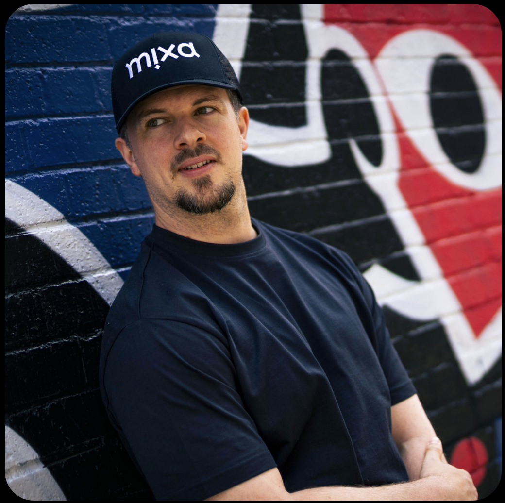 A man wearing a black cap with the word 'mixa' on it, standing against a colorful brick graffiti wall.