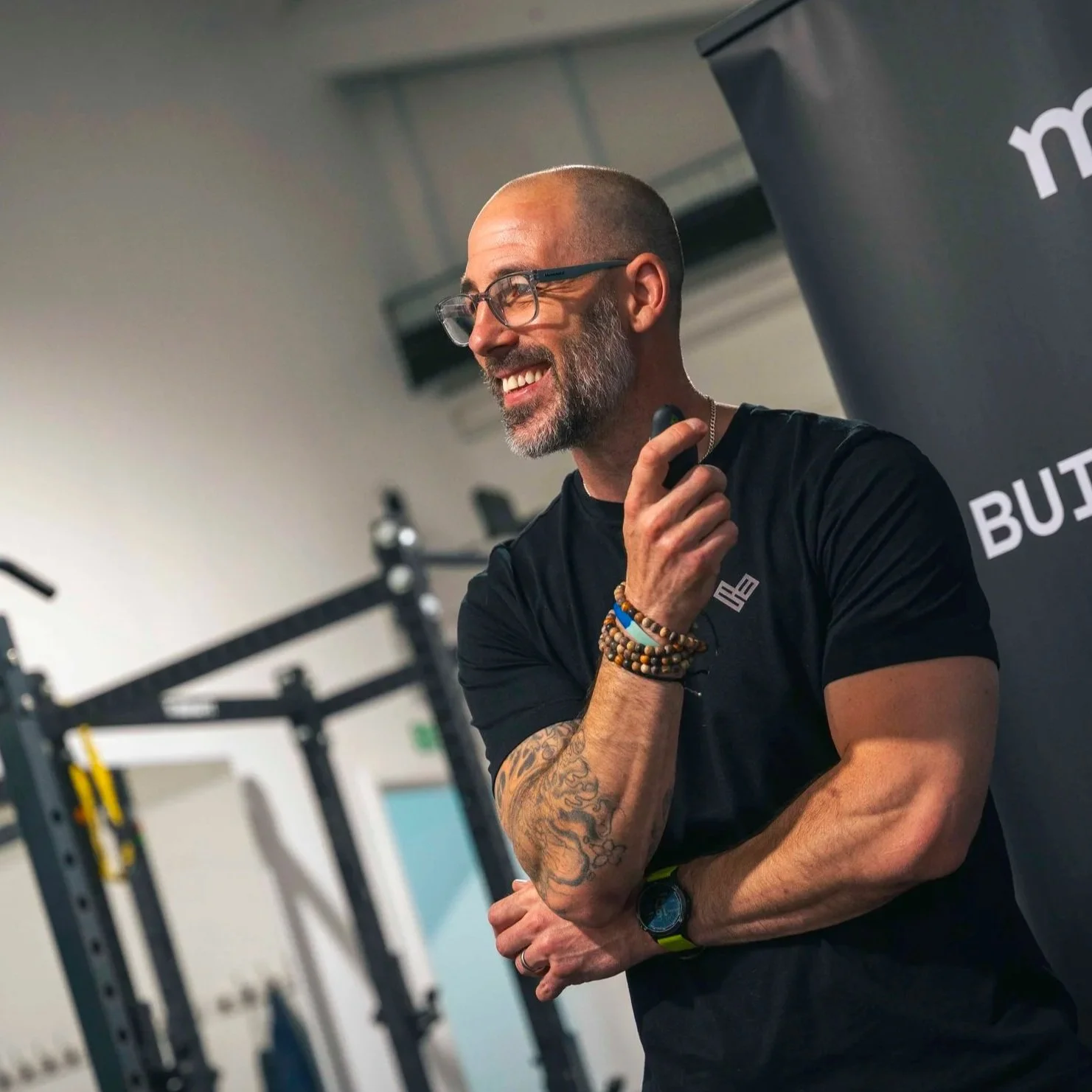 A smiling man with glasses, a beard, and tattoos, wearing a black shirt and colorful bracelets, stands in a gym near workout equipment, holding a microphone.