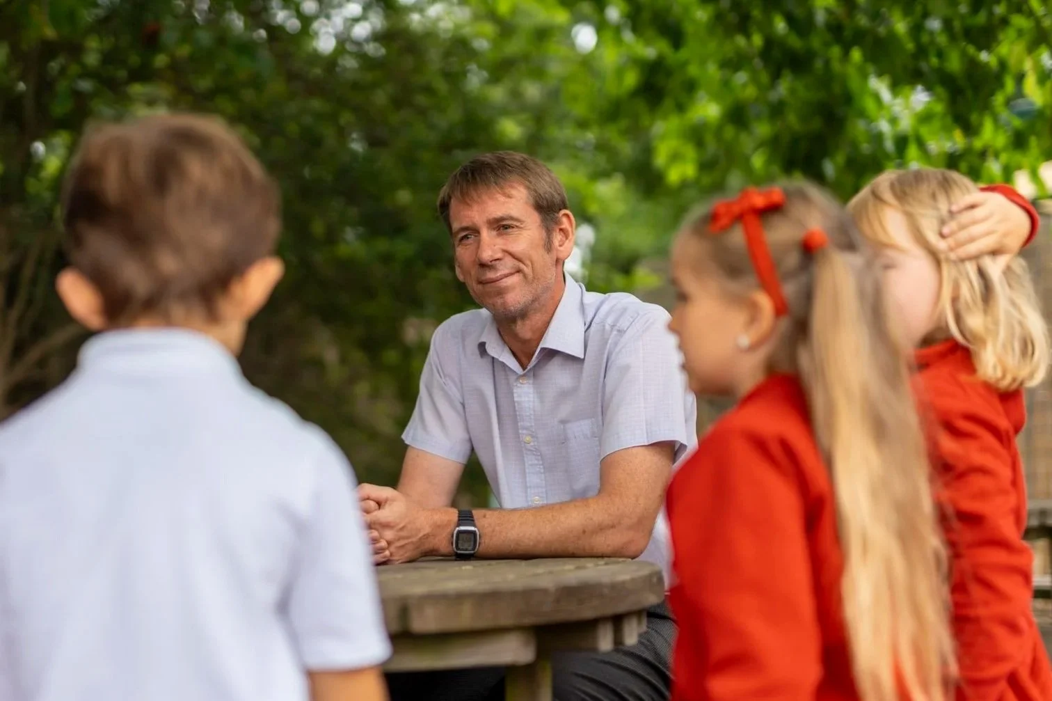 A man in a light blue shirt sitting at a wooden table outdoors having a conversation with three children, two girls and a boy, with green trees in the background.