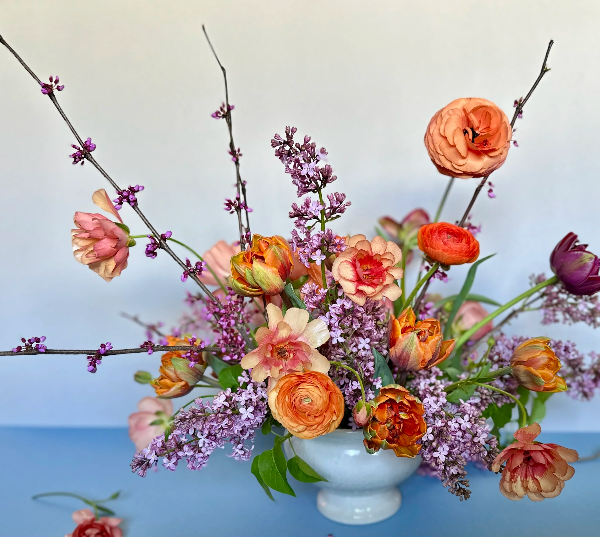 A colorful floral arrangement in a white vase with pink, orange, and purple flowers and green leaves on a blue surface against a plain background. Portland floral designer, Portland wedding florist, Oregon wedding flowers, flowers for delivery.