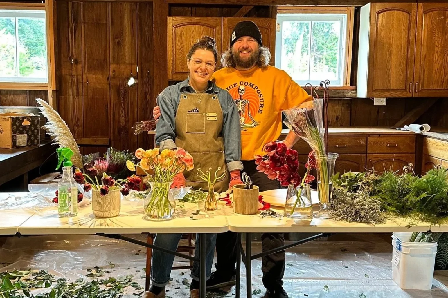 Two people smiling inside a wooden-themed room, standing behind a table with various flowers and floral arrangements, with windows in the background. Portland floral designer, Portland wedding florist, Oregon wedding flowers, flowers for delivery.