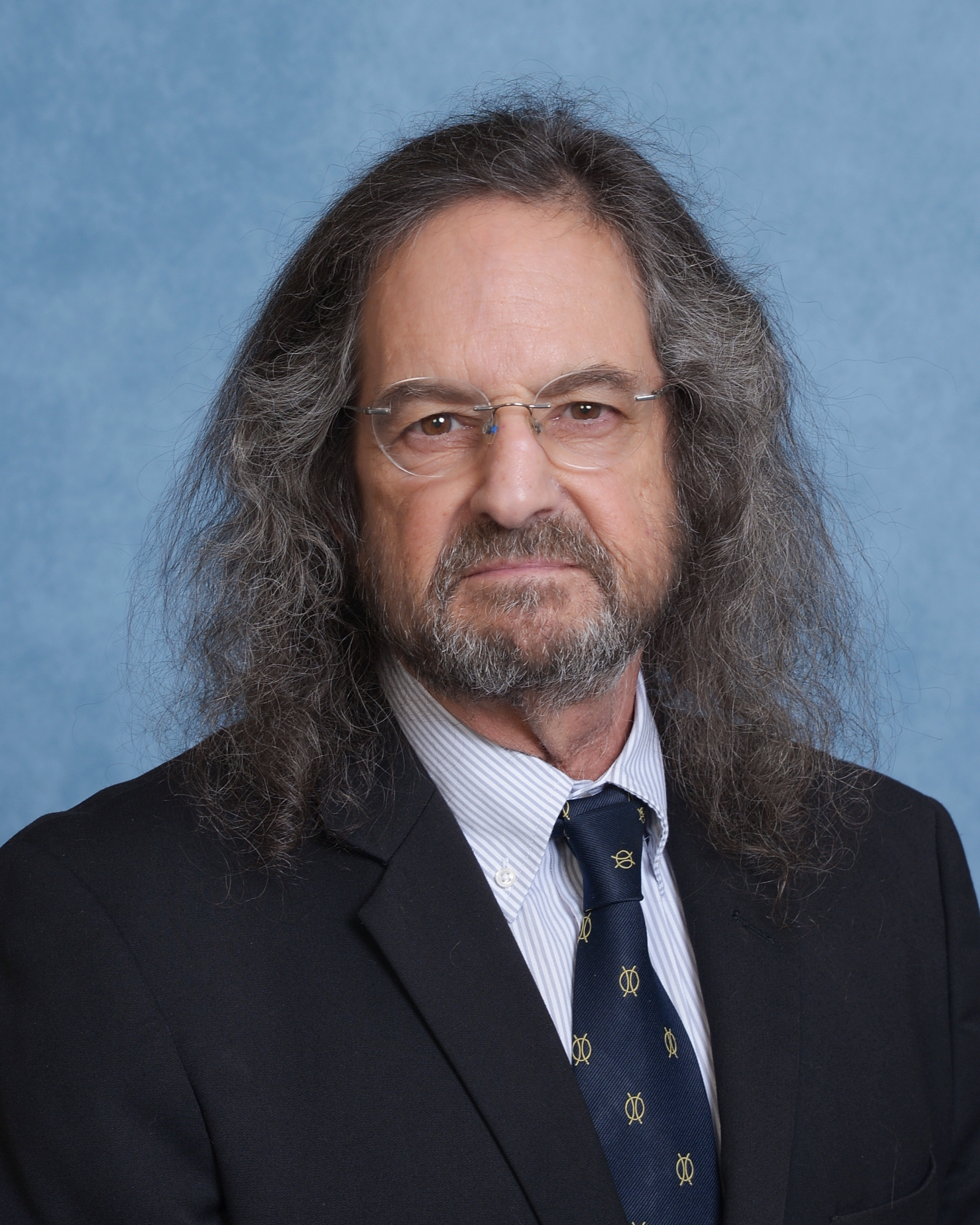 Headshot of Web Sparks wearing a dark suit with a striped shirt and a patterned tie, posing against a blue background.