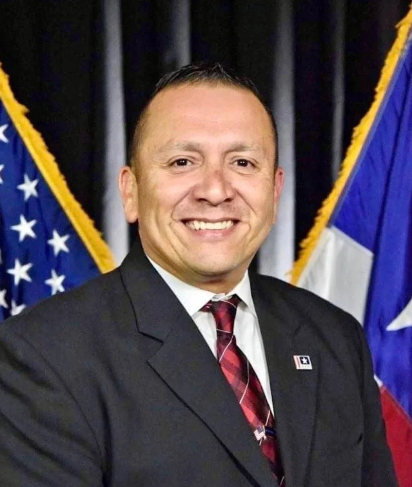 A man in a suit smiling in front of American and Texas flags.