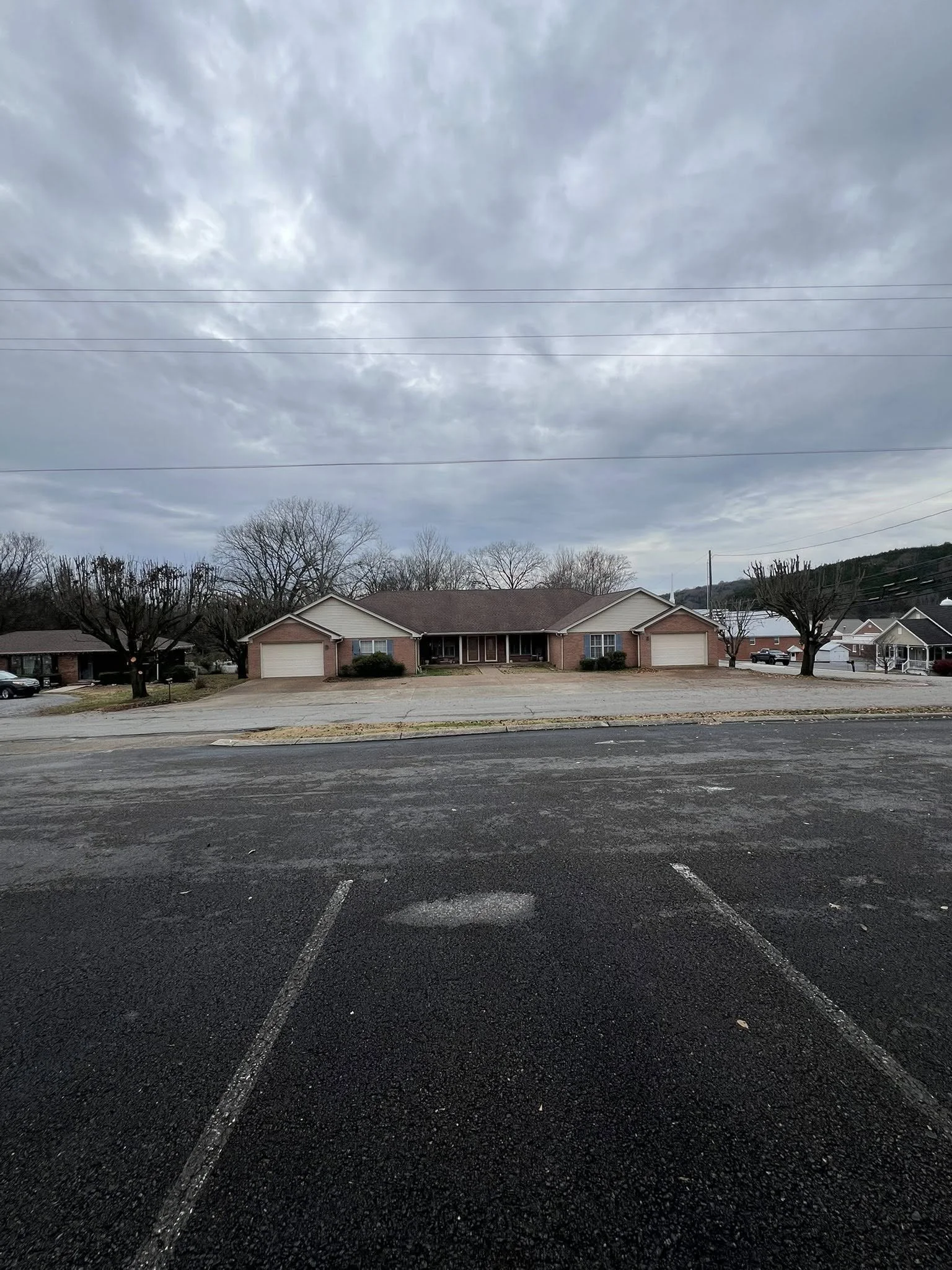 A residential neighborhood with a large brick house with attached garages, trees without leaves, and a cloudy sky. Empty parking lot in foreground.
