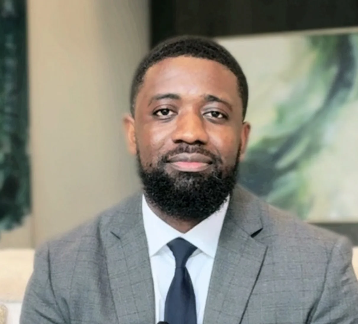 A man in a gray suit with a white shirt and dark tie, with a beard and short hair, posing indoors with a blurred background and a large artwork.