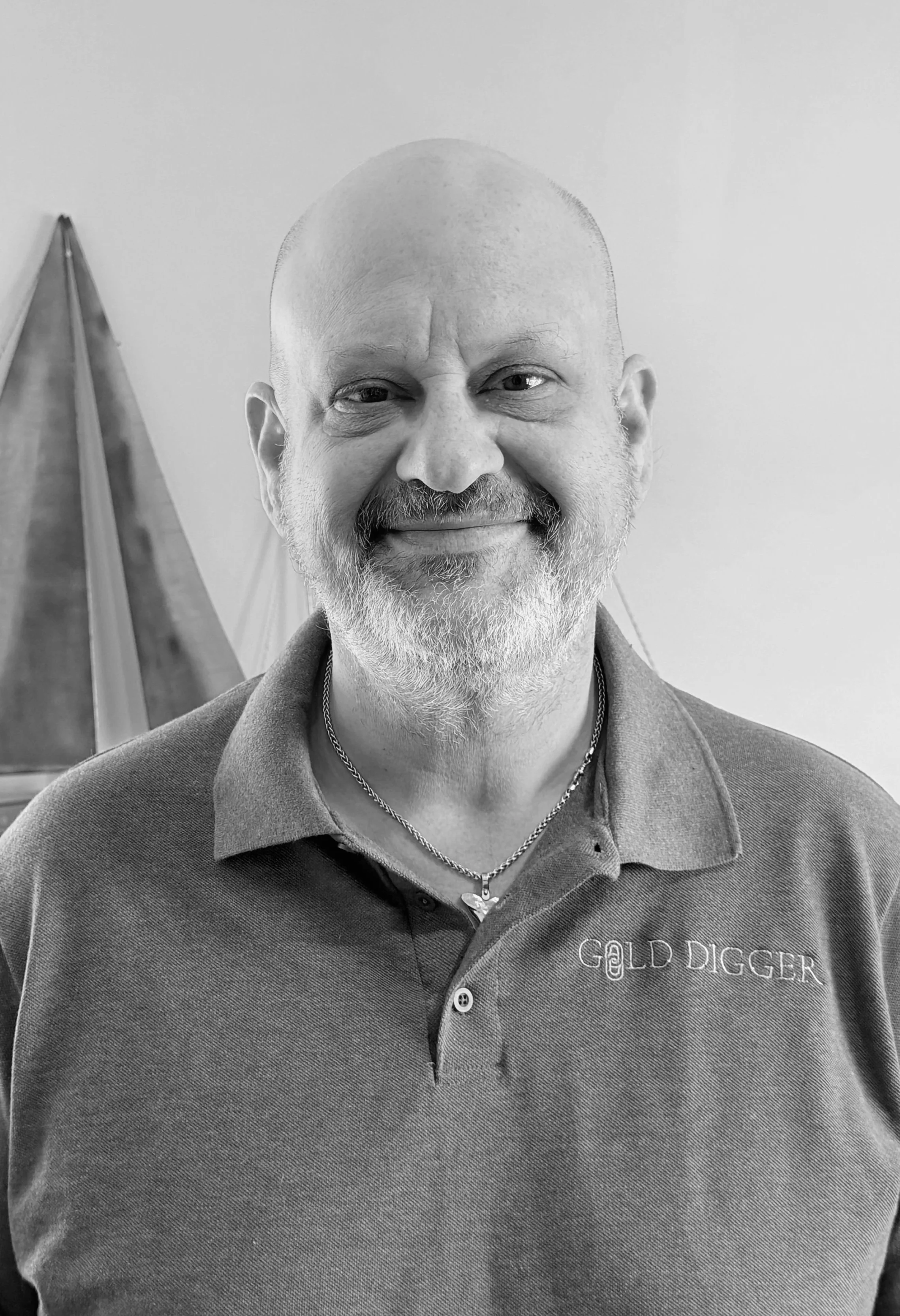 A smiling man with a beard and mustache wearing a polo shirt and necklace, standing indoors.