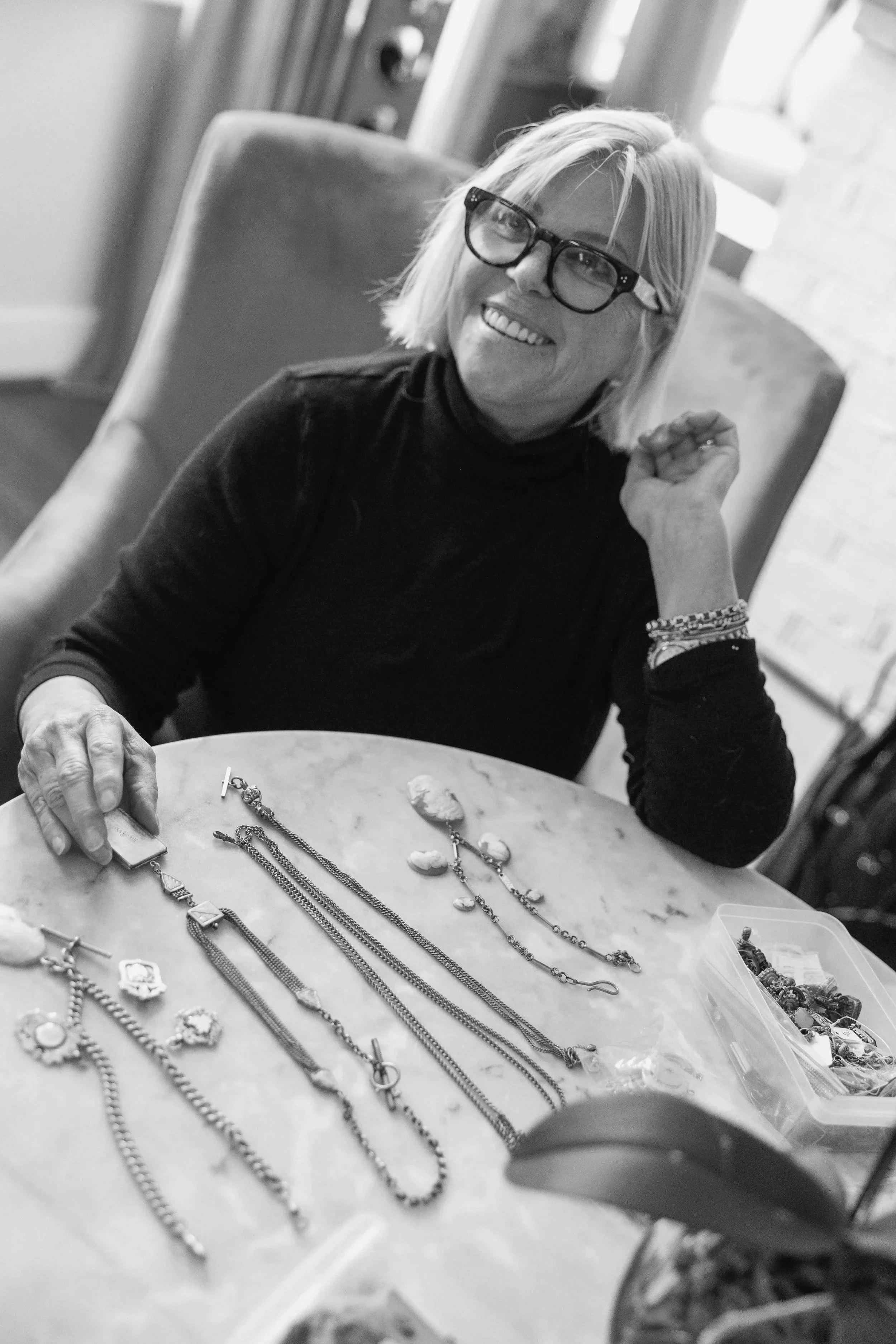 A woman with glasses sitting at a table with jewelry, smiling and looking to her left.