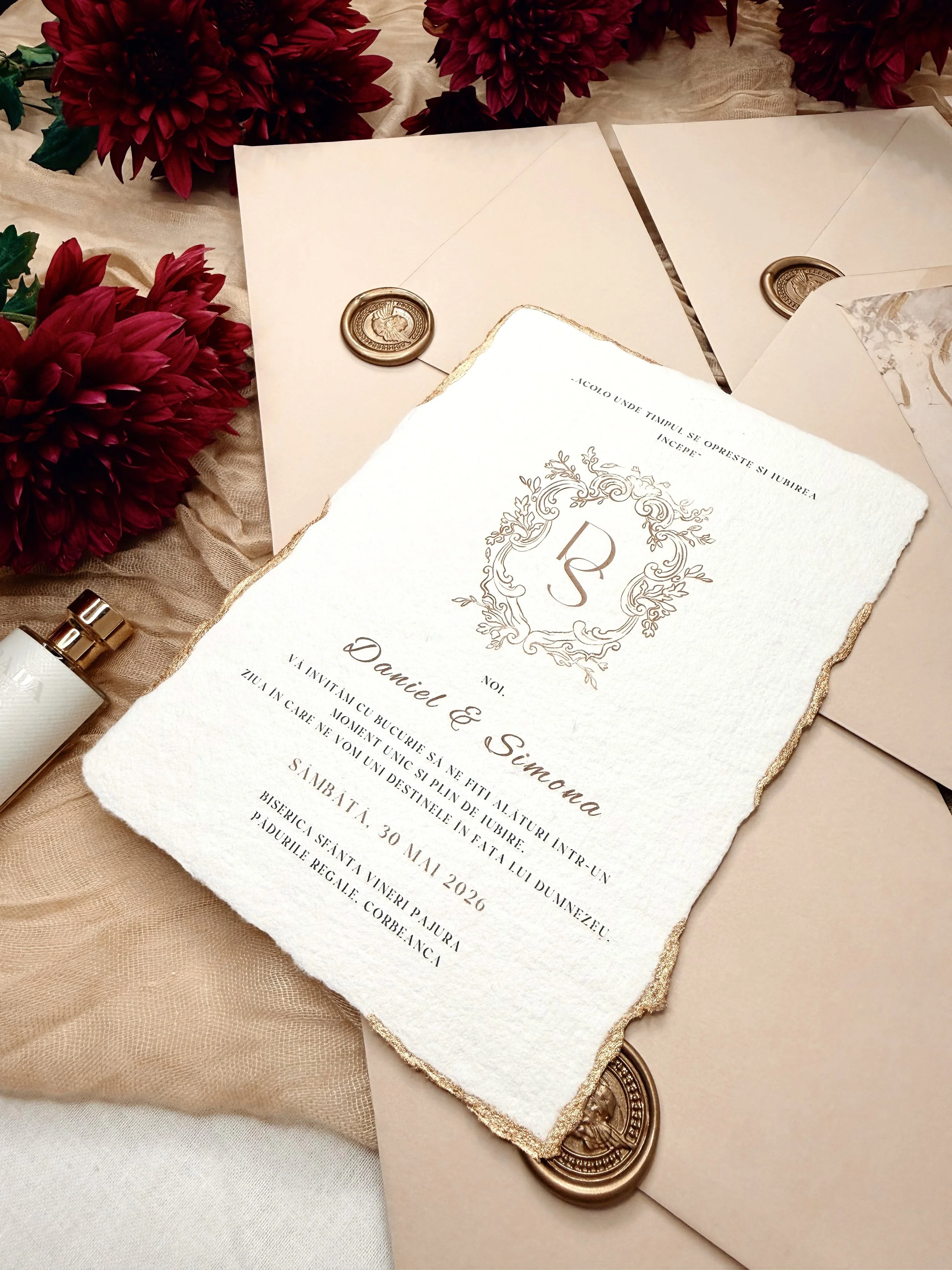 Wedding invitation with ornate border and monogram, surrounded by pink flowers, beige fabric, sealed envelopes with gold wax seals, and a white pen.