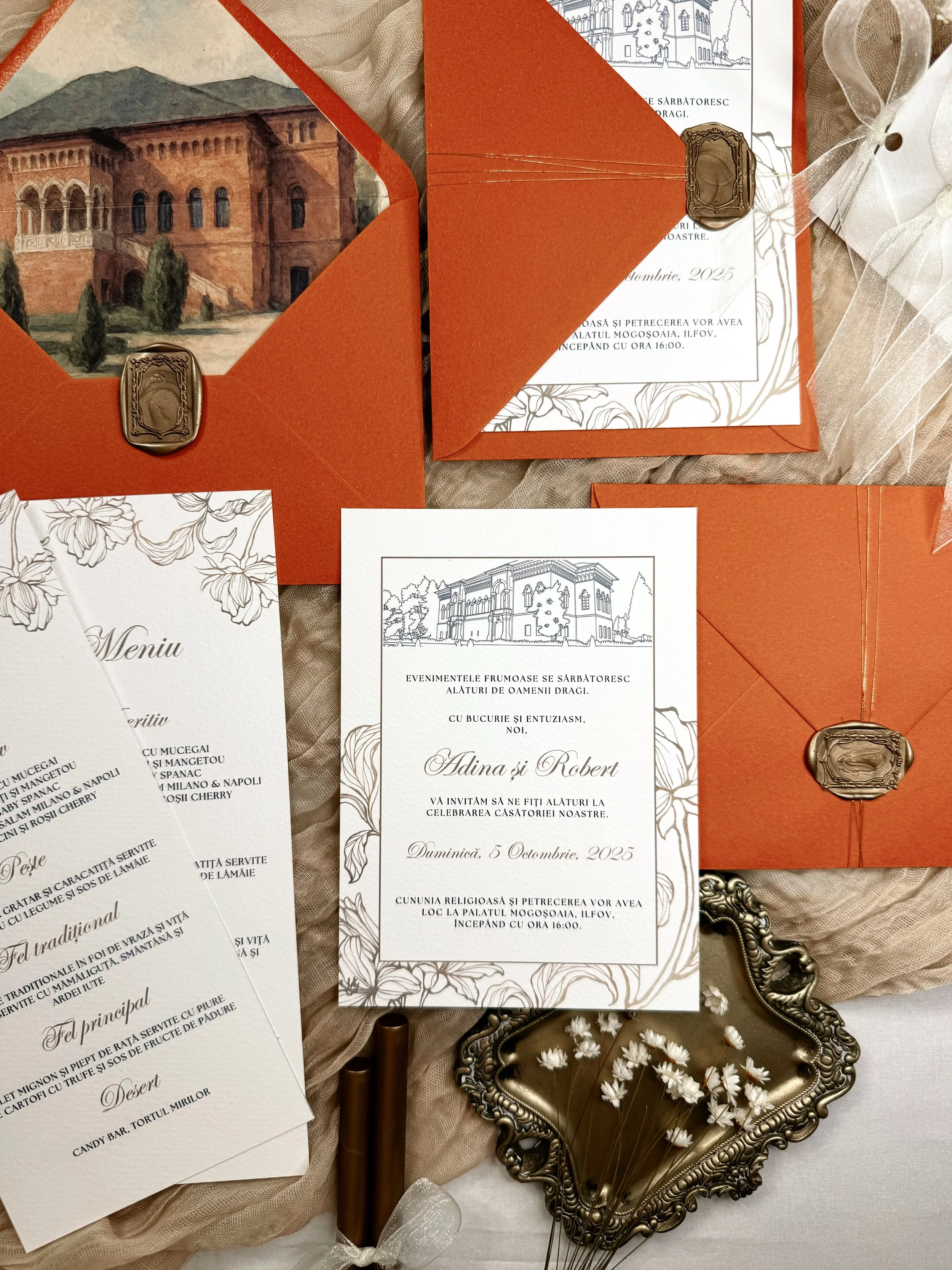 Wedding invitation set and decor featuring orange envelopes with wax seals, printed menus and invitation cards with floral and architectural designs, vintage-style mirror tray with white flowers, black pens, and decorative elements on fabric background.