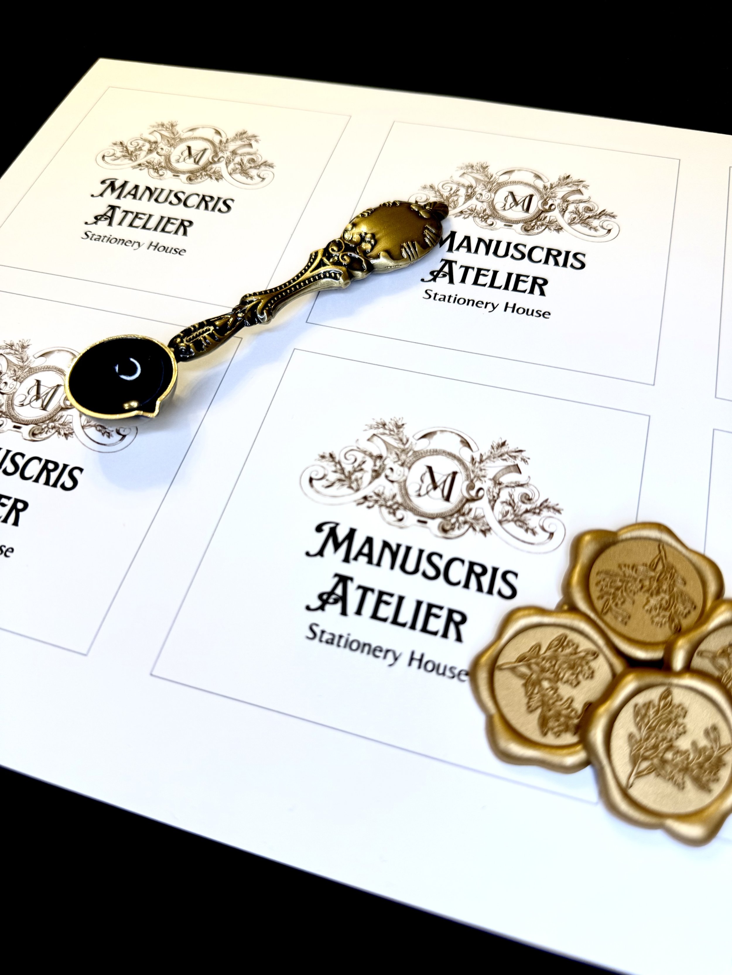 Four sheets of paper with 'MANUSCRIS ATELIER Stationery House' written on them, decorated with ornate designs and an 'M' logo. Several gold seals with floral imprints and an ornate gold-handled wax seal stamp are placed on top of the sheets.
