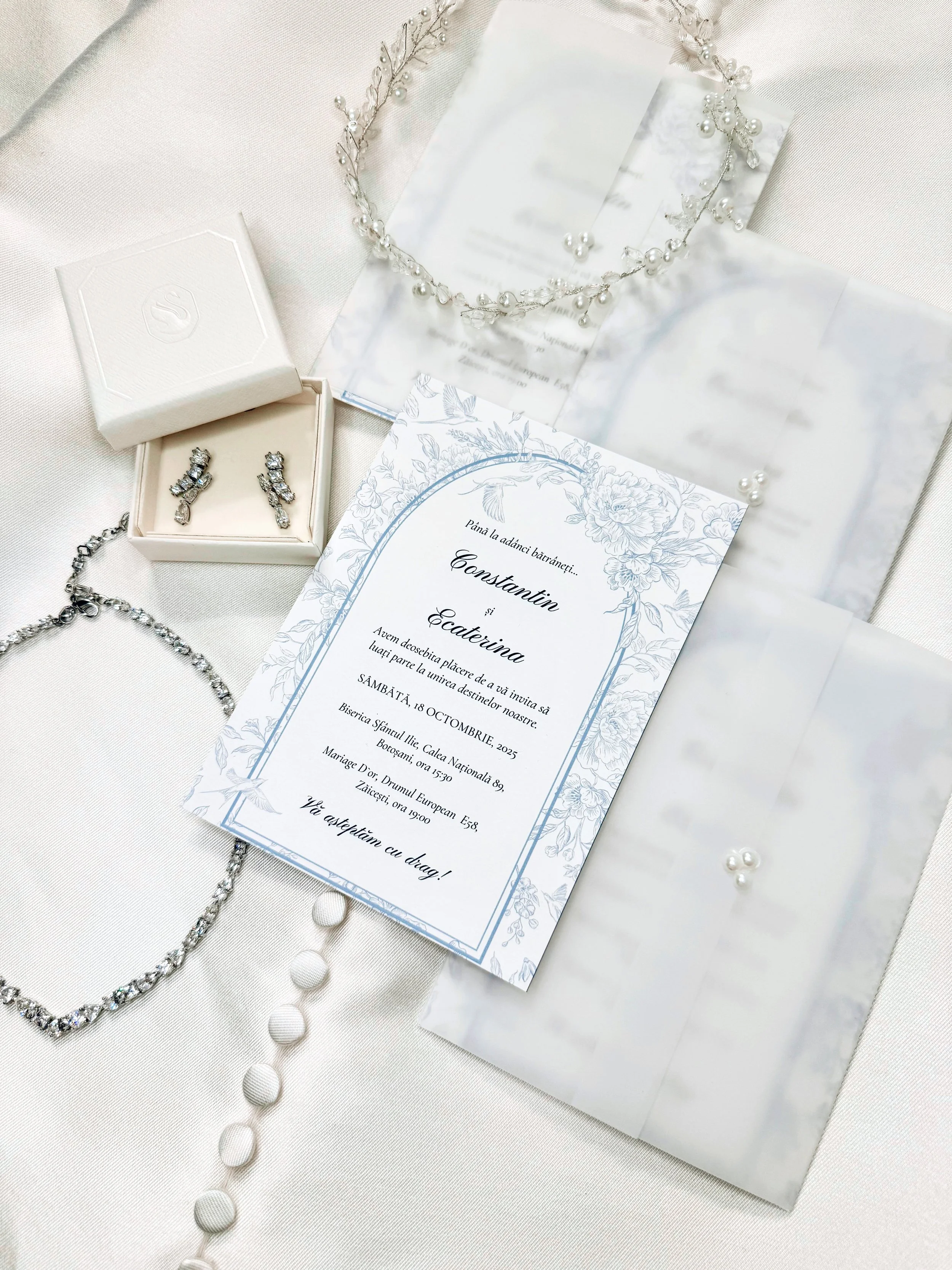 Wedding invitation card with floral design, jewelry including earrings, necklaces, and a pearl headband, on a white fabric background.