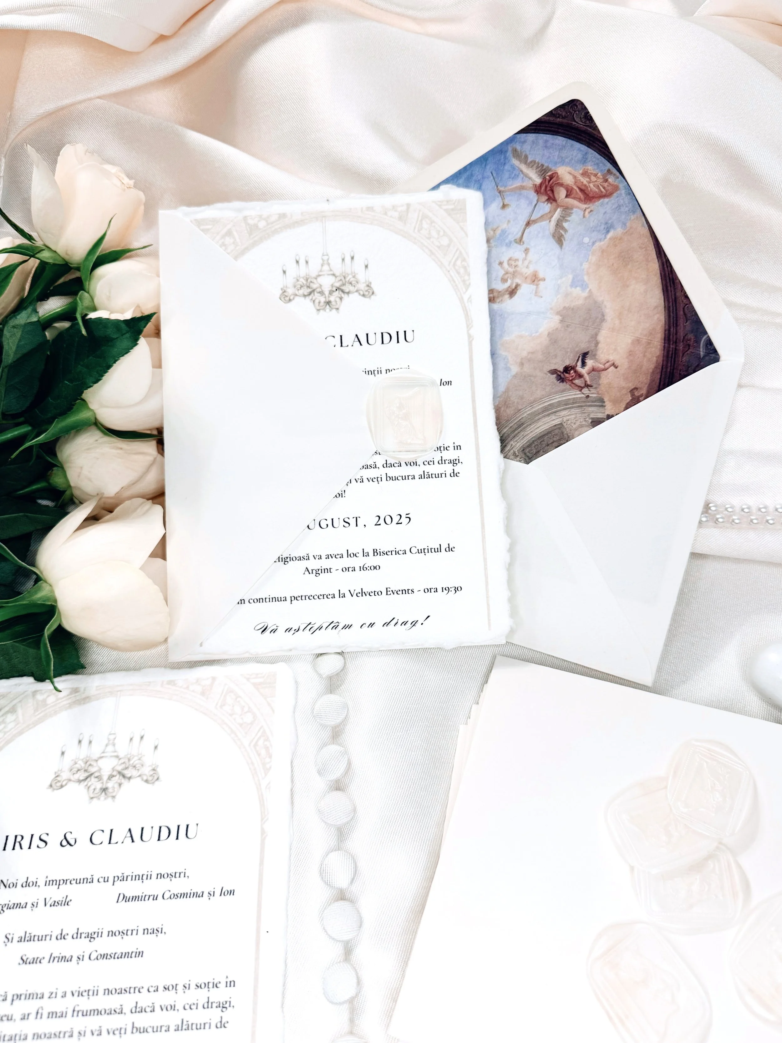 Wedding invitation, a bouquet of white roses, and an open card with a mural painting inside, all set on white fabric.