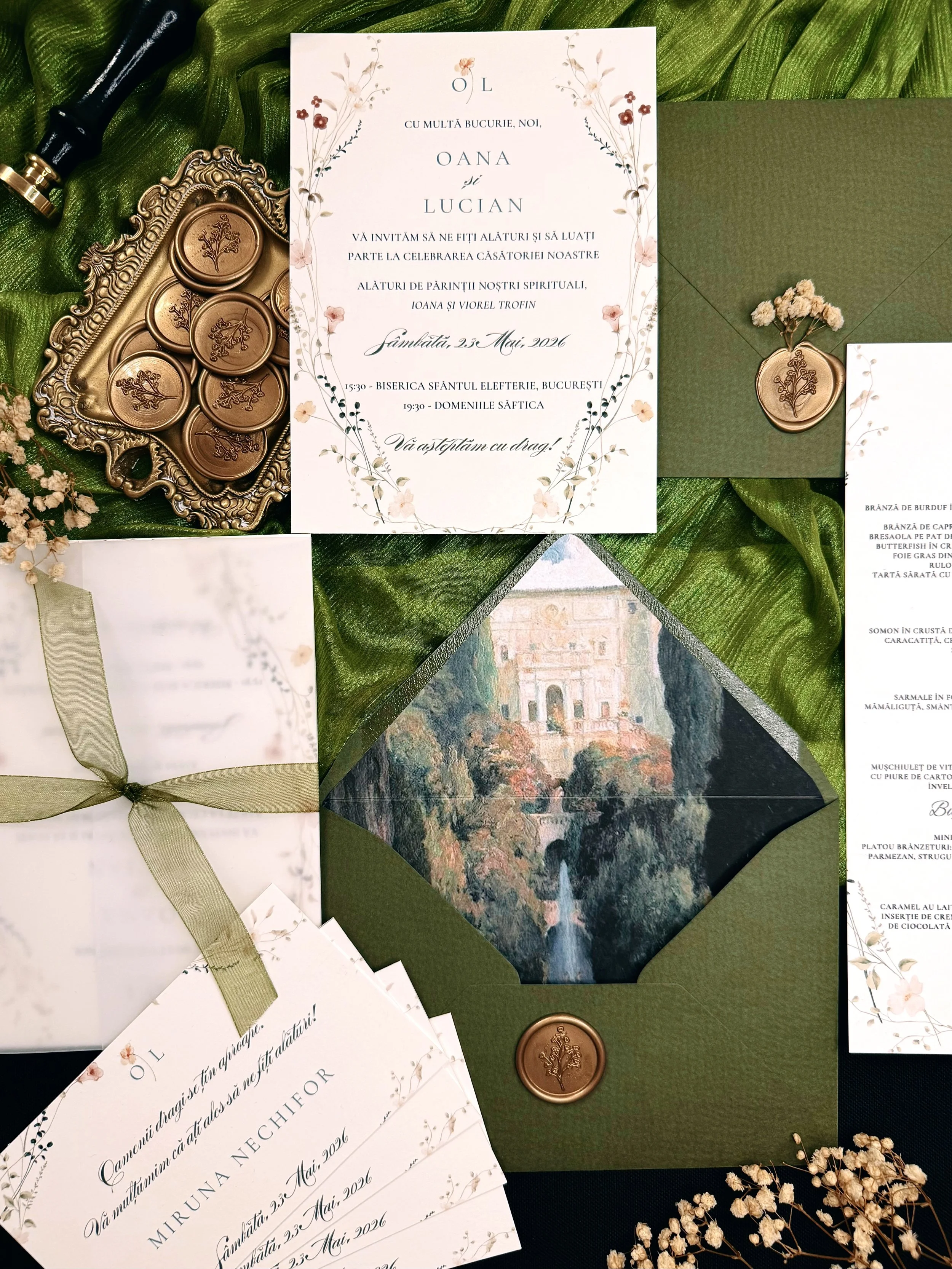 Invitation card with floral design and wedding details, surrounded by wax seals, a floral illustration, and a gift box with a ribbon, all placed on a green fabric background.